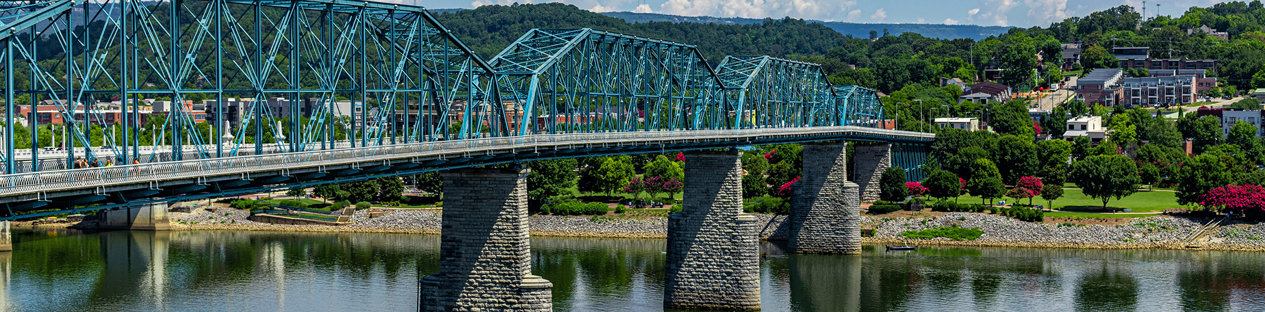 Chattanooga bridge crossing the river with green trees and buildings in the background under a blue sky.