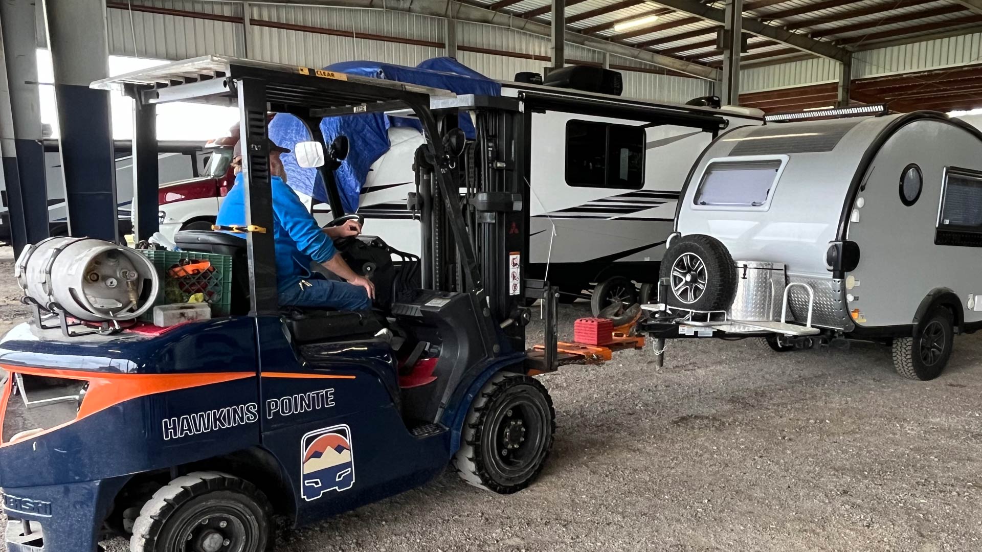Staff operating a Hawkins Pointe forklift inside a warehouse, lifting a small silver camper trailer.