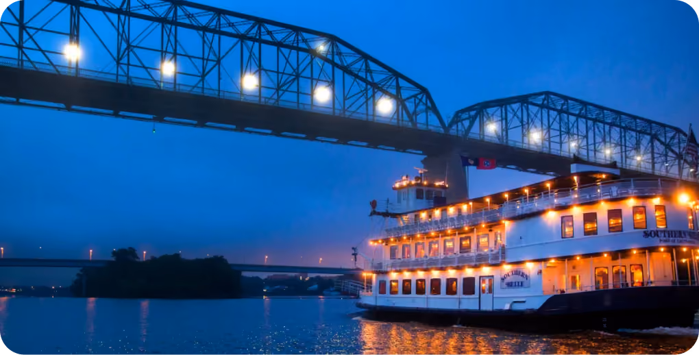 Illuminated Southern Belle Riverboat cruising under a bridge at night.