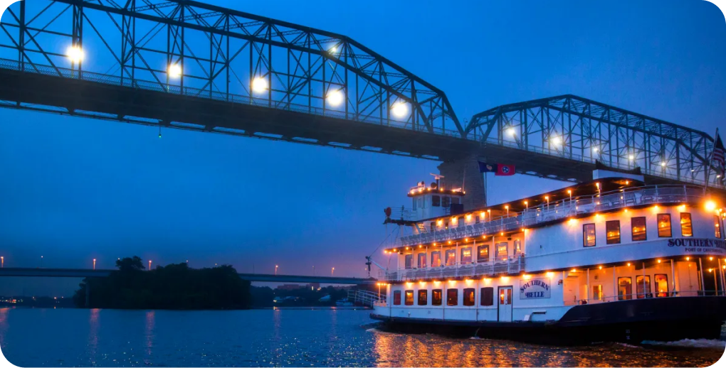Illuminated Southern Belle Riverboat cruising under a bridge at night.