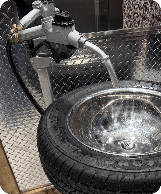 Restroom sink built from a Firestone tire with a gas pump nozzle faucet against diamond plate walls.