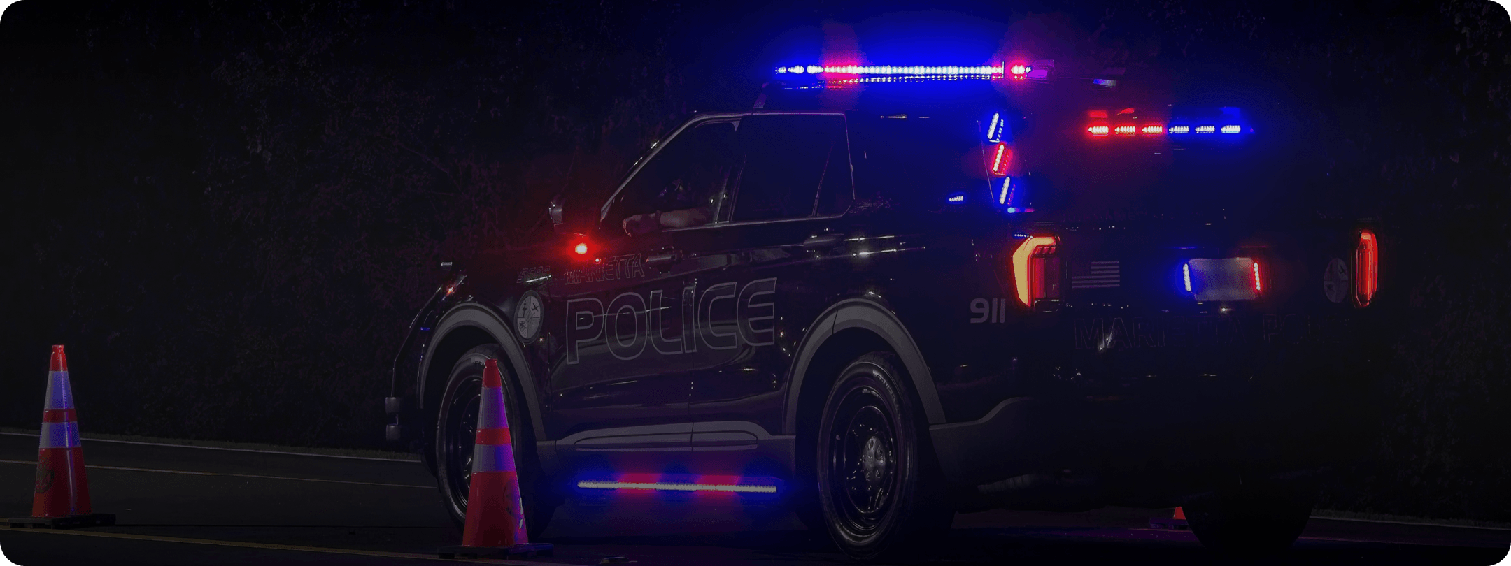 Police SUV parked at night with red and blue emergency lights flashing next to orange traffic cones.