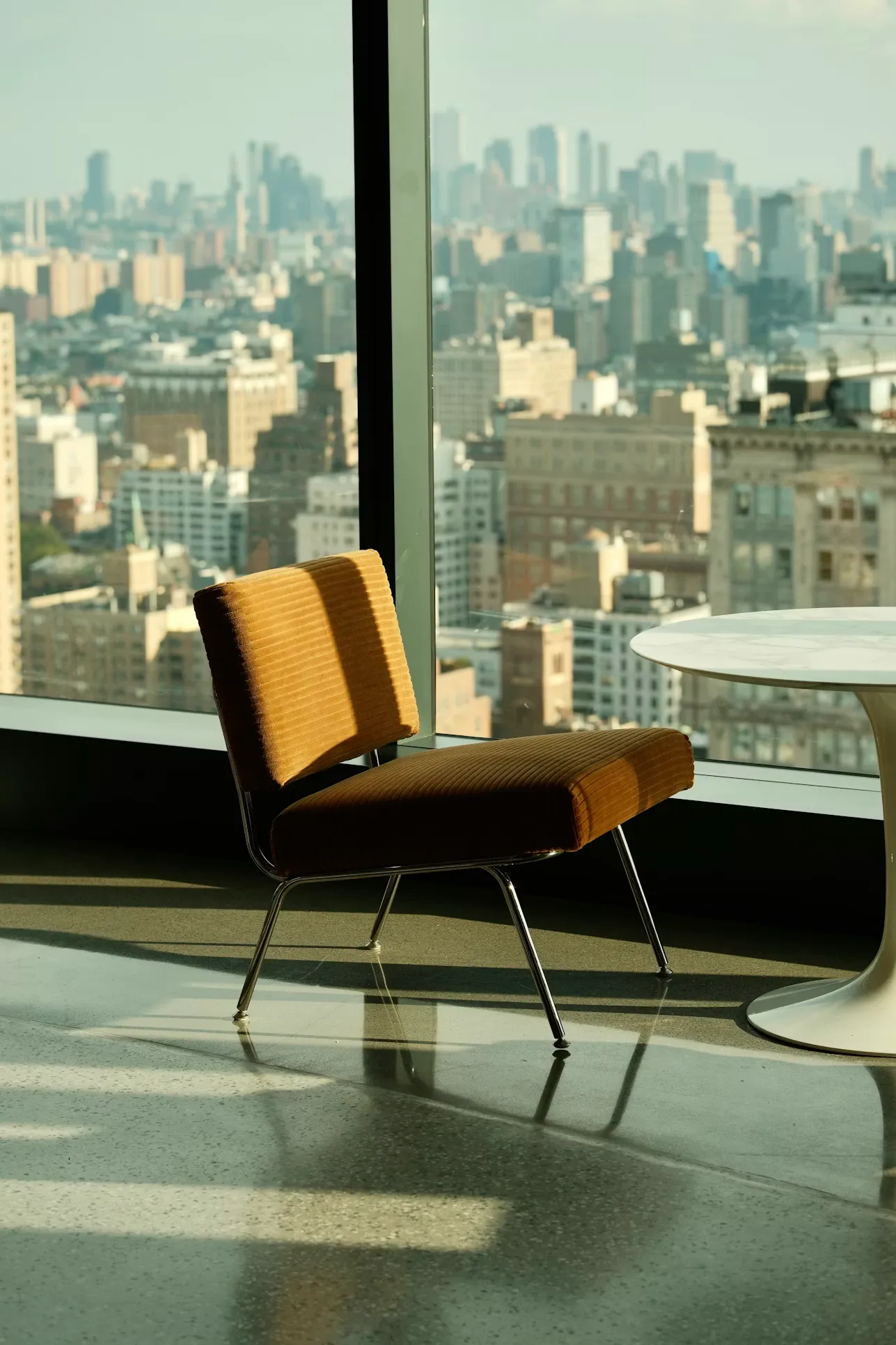 Brown cushioned chair near a white round table by a large window with a city skyline view.