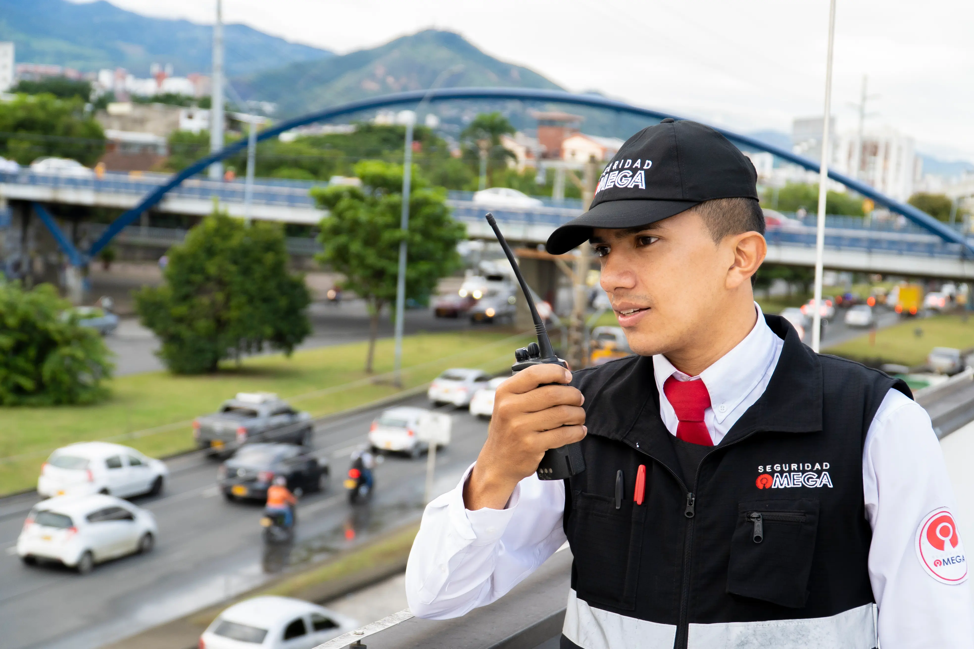 Security guard in black vest and cap using a walkie-talkie overlooking a busy urban road with cars and motorcycles.