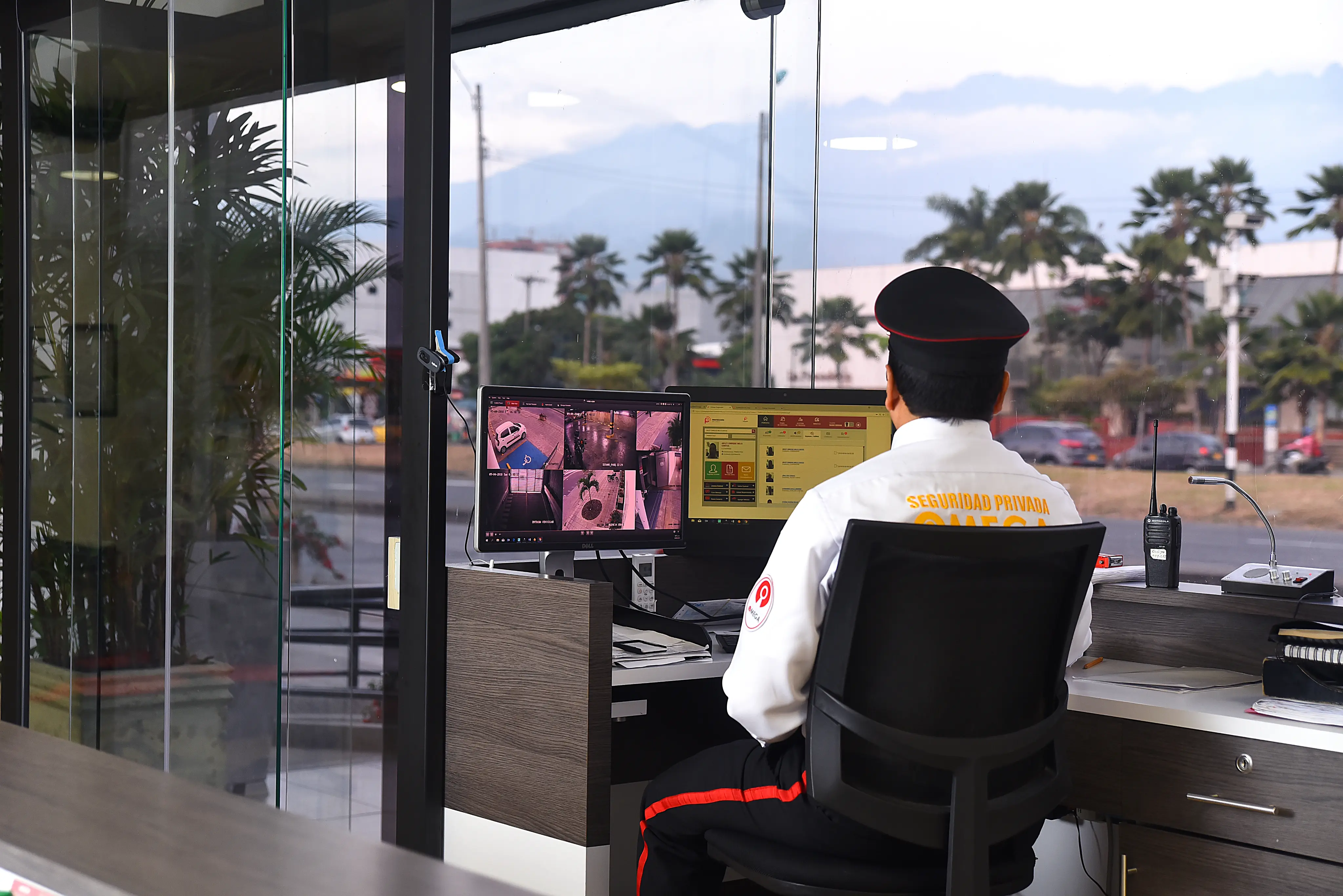 Security guard in uniform monitoring multiple surveillance camera feeds on two computer screens inside a glass-walled office.