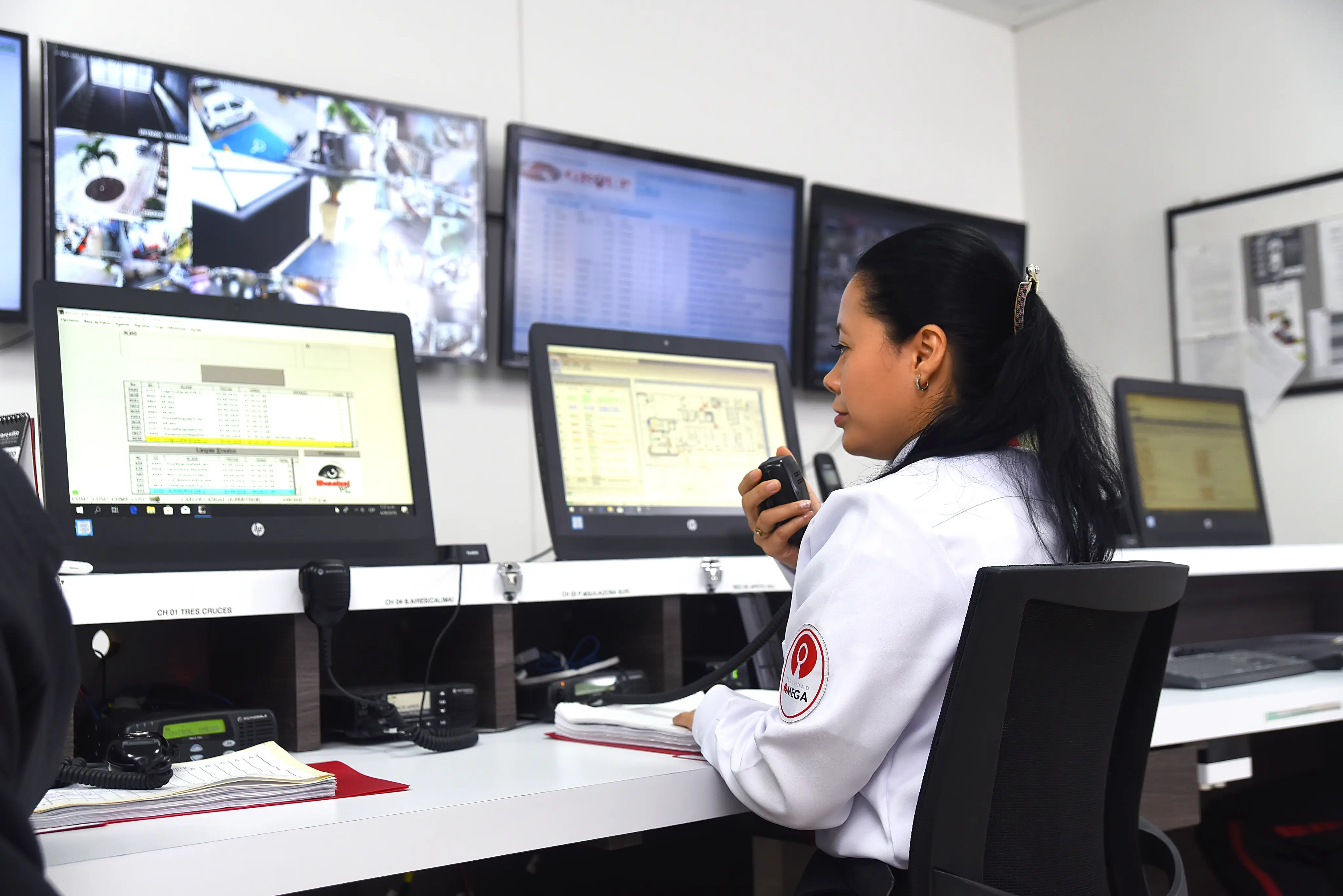 Woman in a security control room speaking into a radio with multiple monitors displaying surveillance footage and data.
