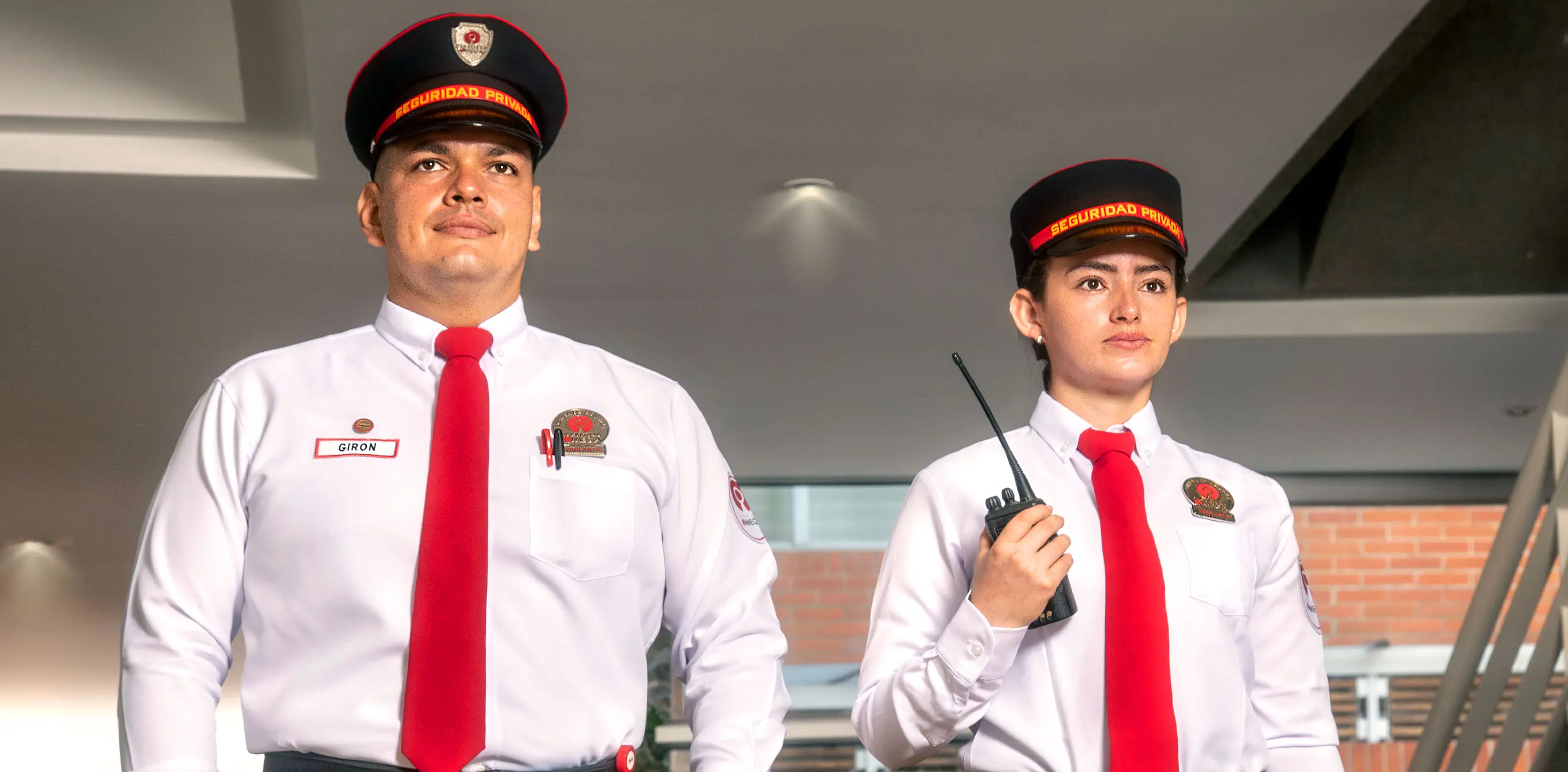 Two security guards in white shirts with red ties and black hats, one holding a walkie-talkie, standing indoors.