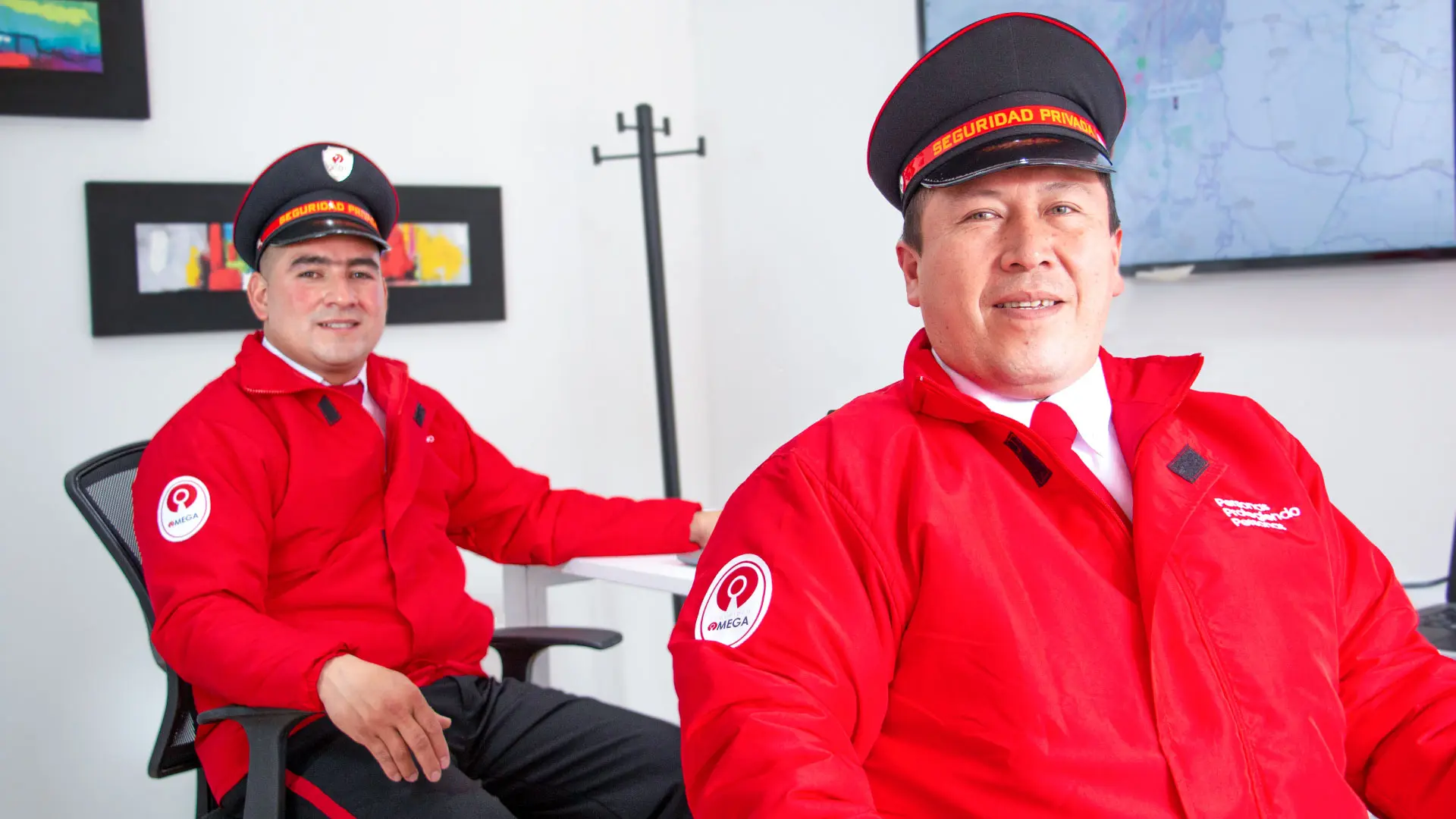 Two men in red security uniforms with hats sitting in an office with white walls and a mounted screen.