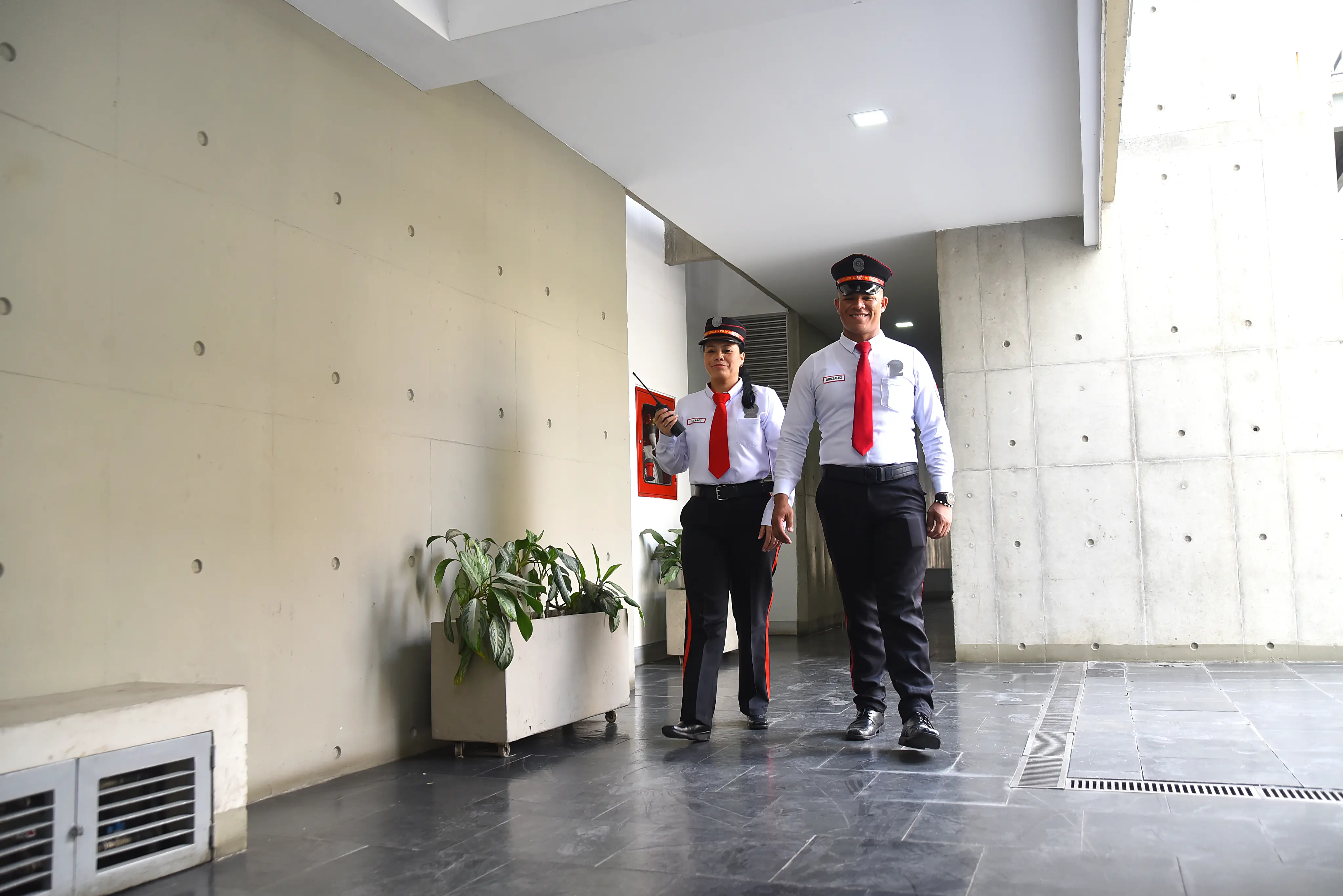 Two security guards walking in a modern indoor corridor with concrete walls and potted plants.