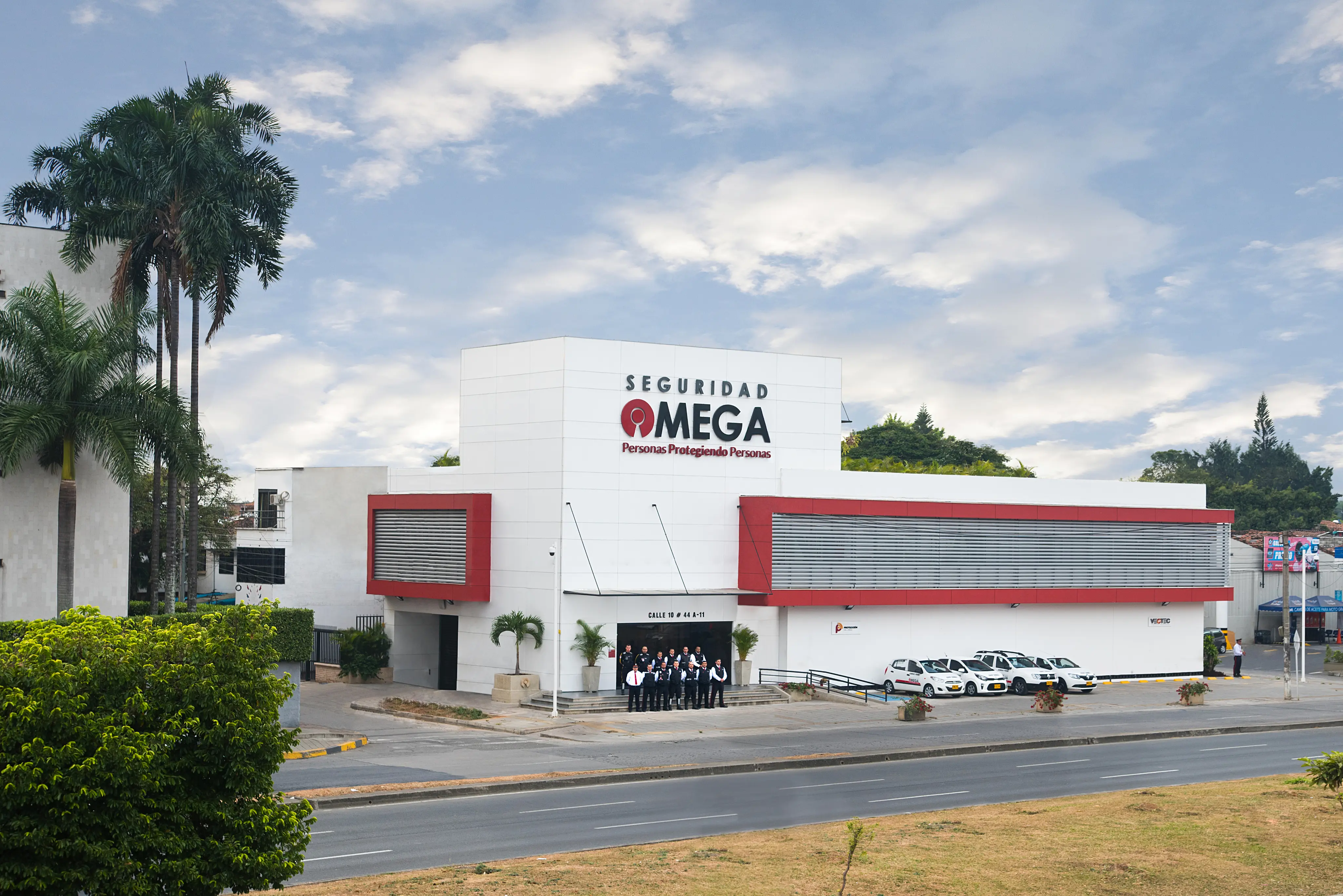 Group of security personnel standing in front of a modern white building with red accents labeled Seguridad Omega and several parked white vehicles.