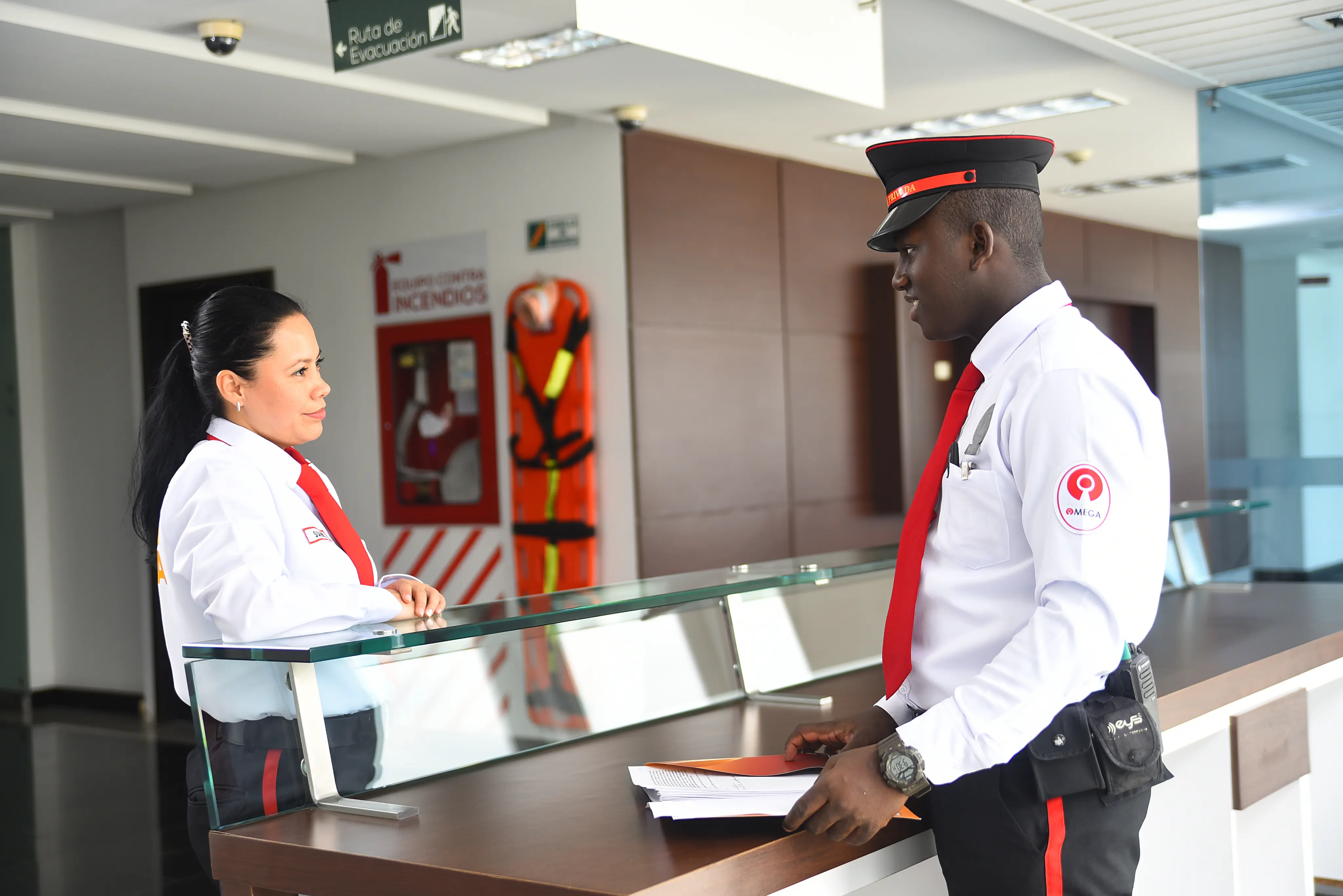 Two security guards in white shirts and red ties standing at a reception desk in a building lobby.