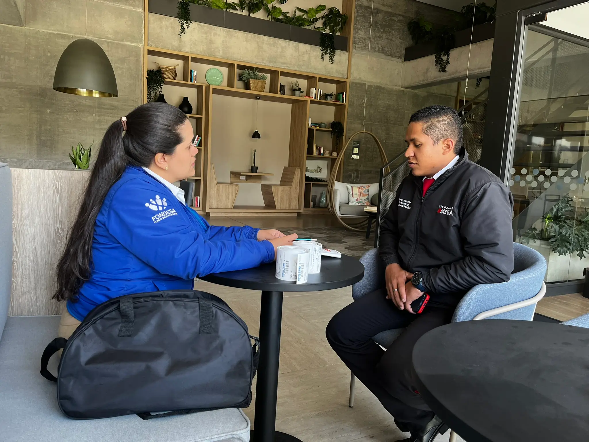 Two people sitting and talking across a small round table in a modern office lounge with plants and shelves in the background.