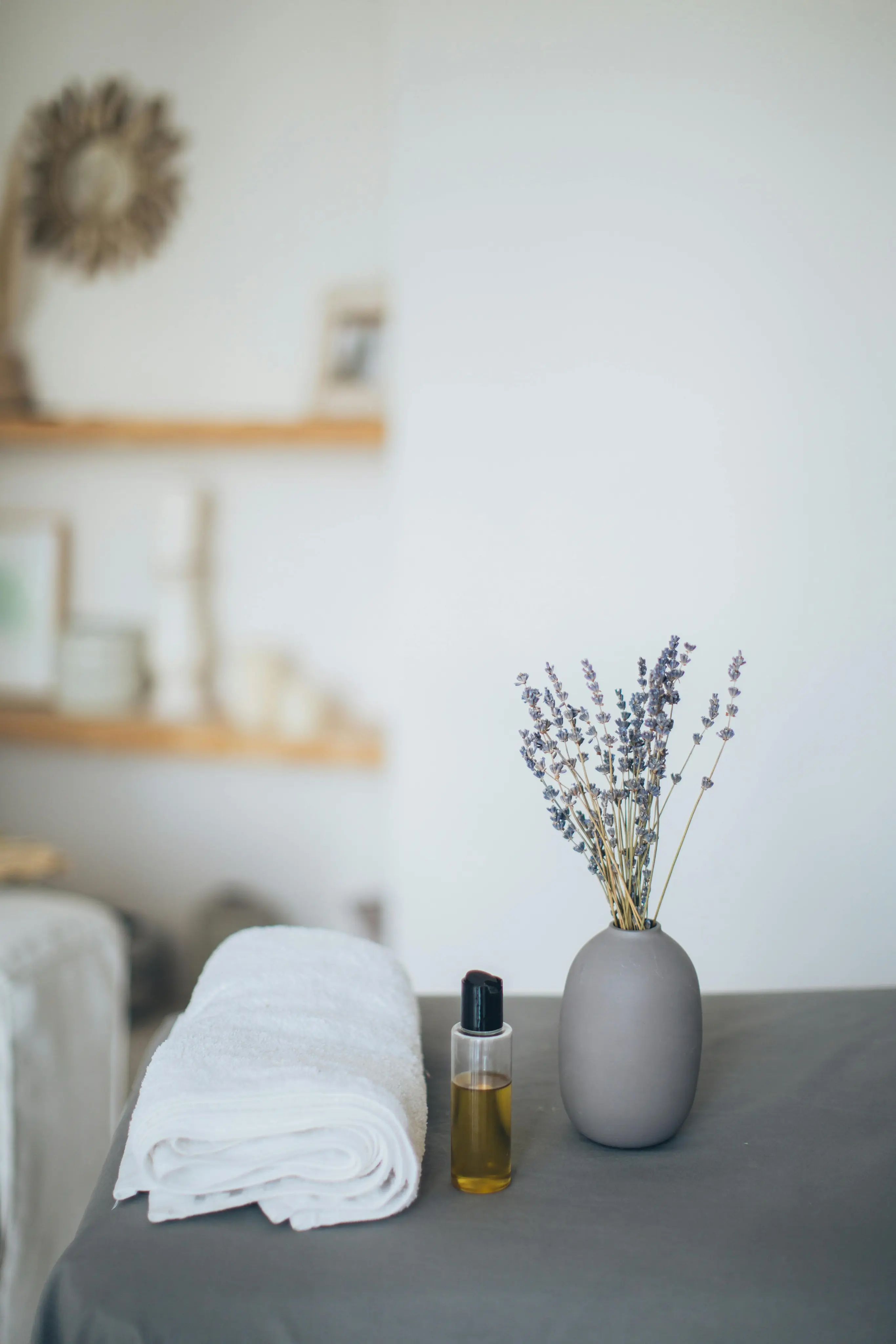Rolled white towel, small bottle of oil, and gray vase with dried lavender flowers on a gray surface in a softly lit room.