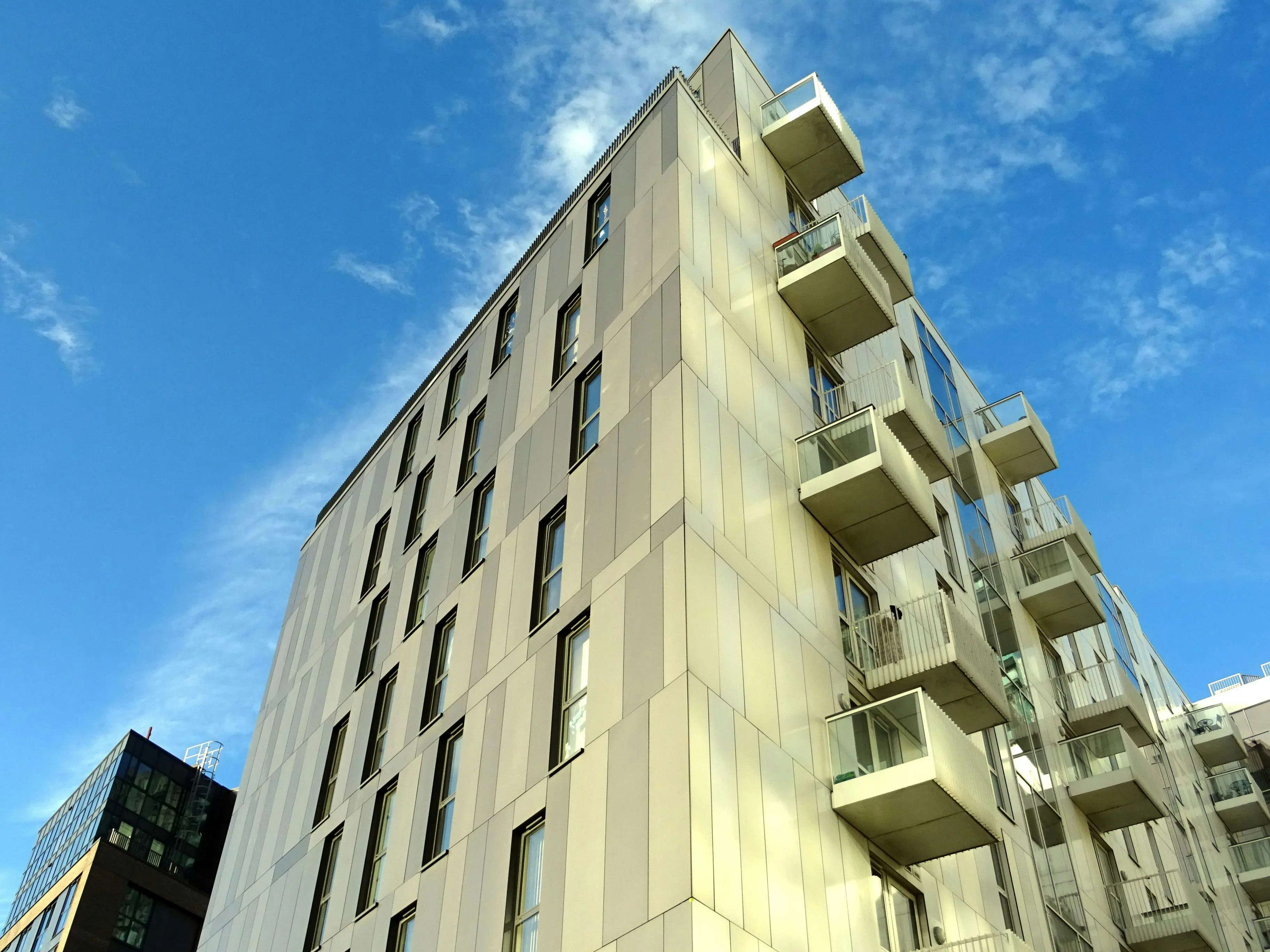 Modern multi-story residential building with beige panels and protruding balconies against a blue sky.