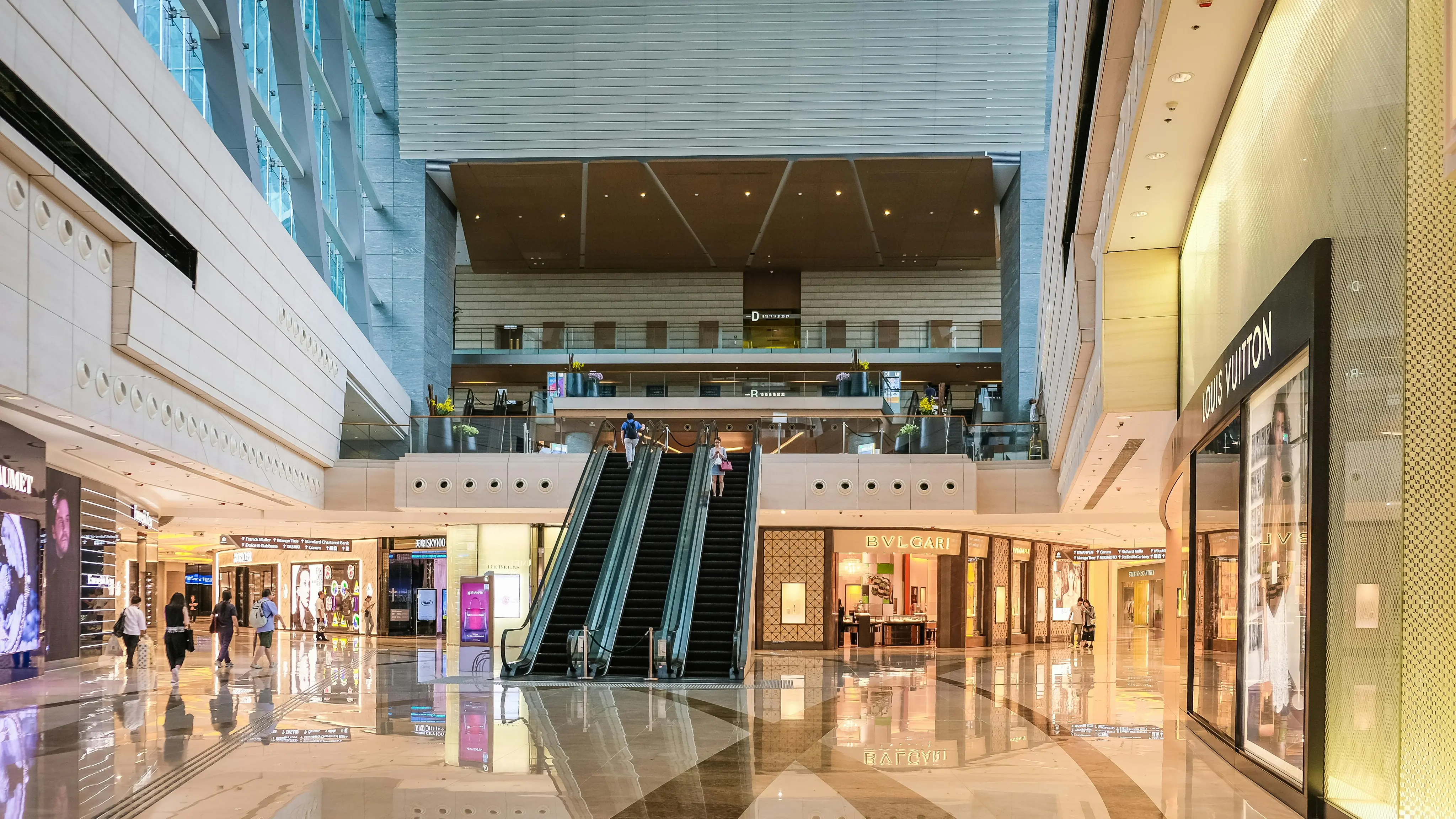 Interior of a luxury shopping mall with escalators in the center and stores like Louis Vuitton and Bvlgari on either side.