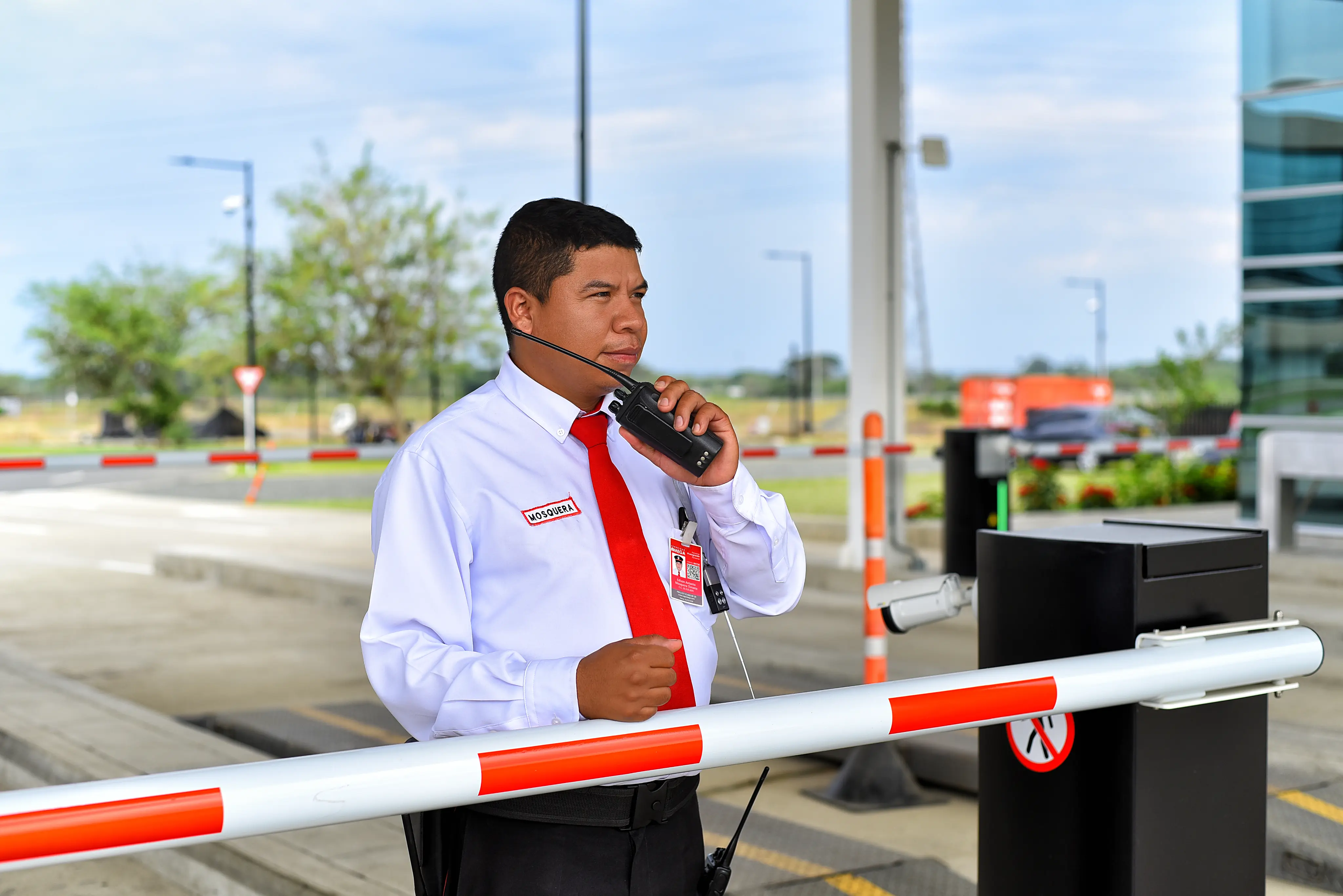 Security guard in white shirt and red tie speaking into a walkie-talkie at a gated entrance.