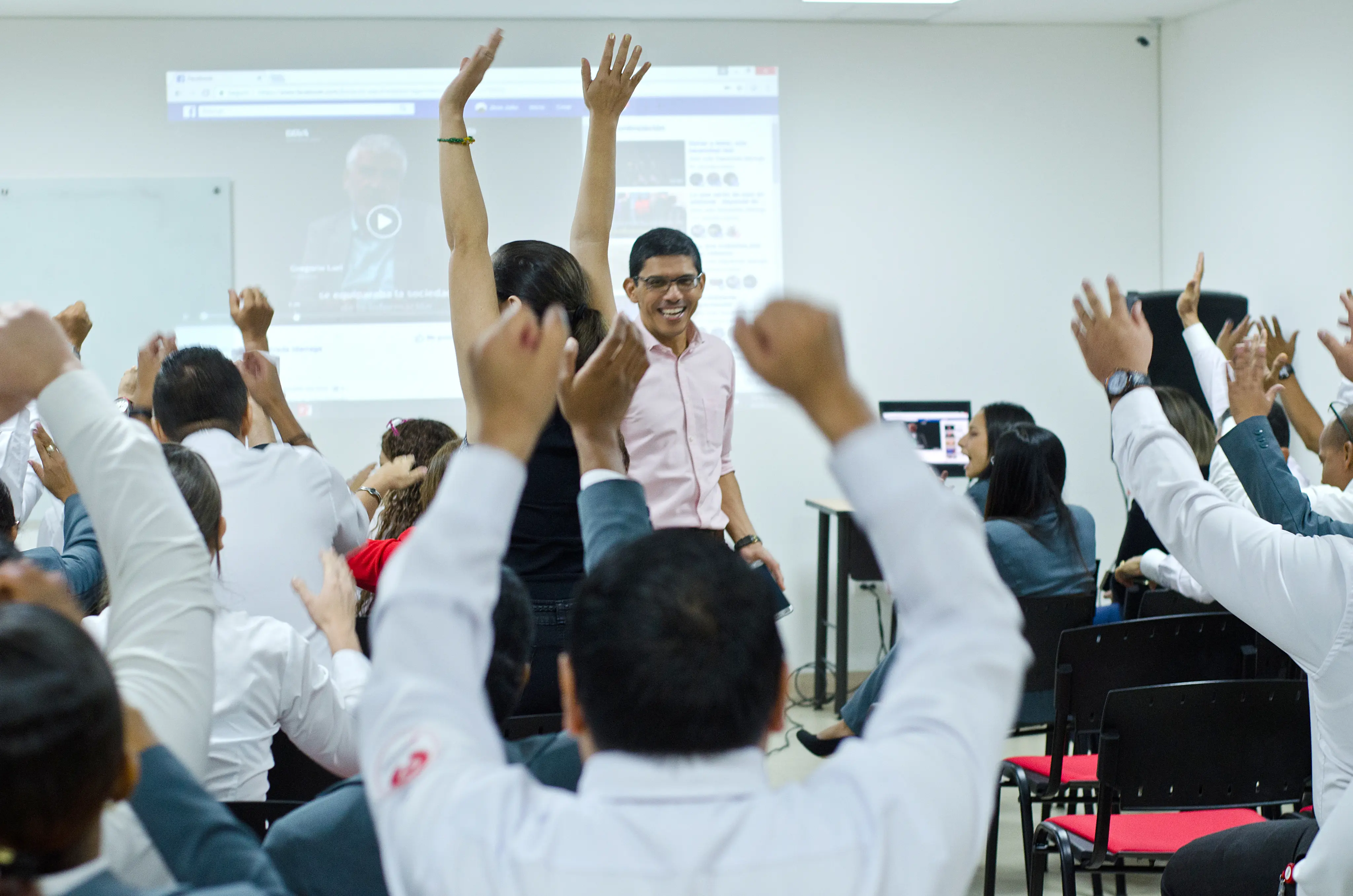 A man in a pink shirt stands smiling in front of a cheering audience with raised hands in a conference room.