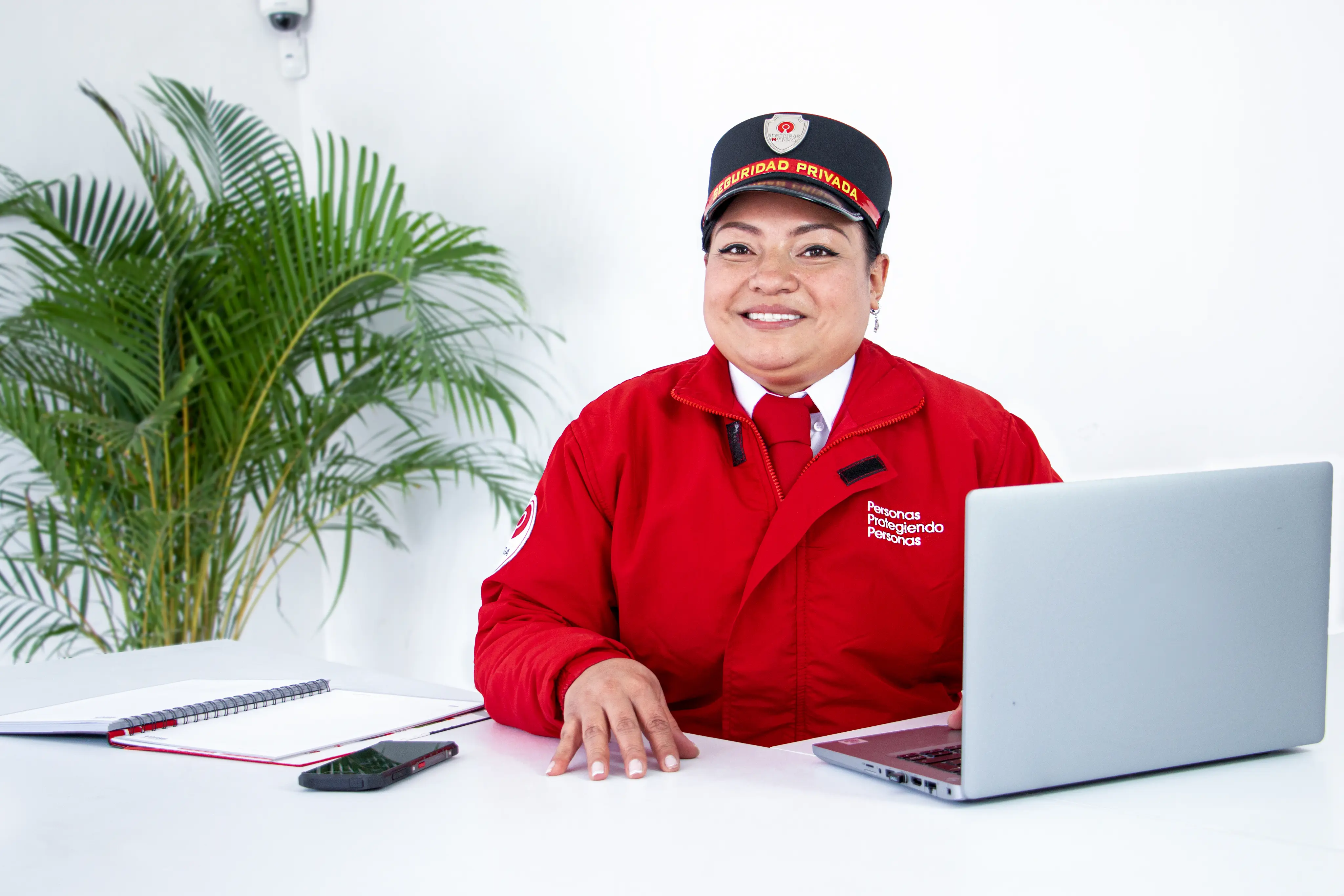Smiling female security guard in a red uniform sitting at a desk with a laptop, notebook, and phone.