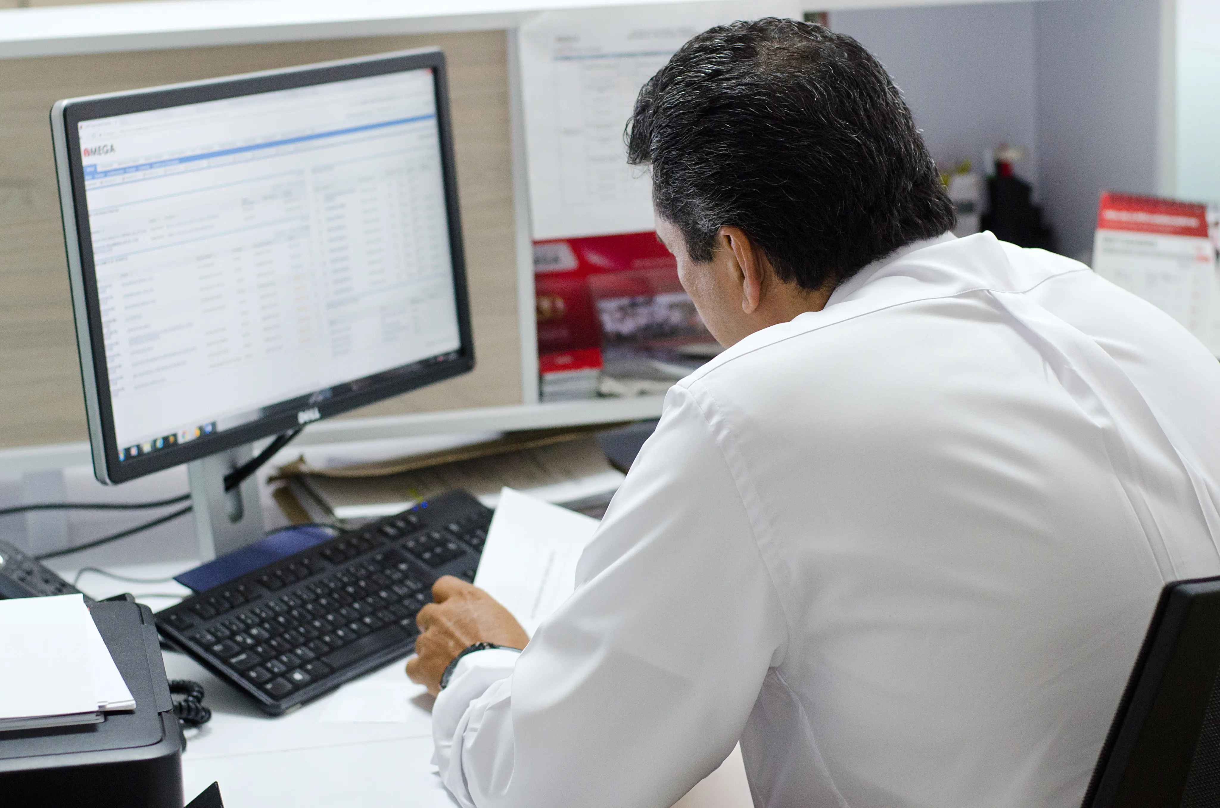 Man in white shirt sitting at a desk, looking at papers with a computer monitor and keyboard in front of him.