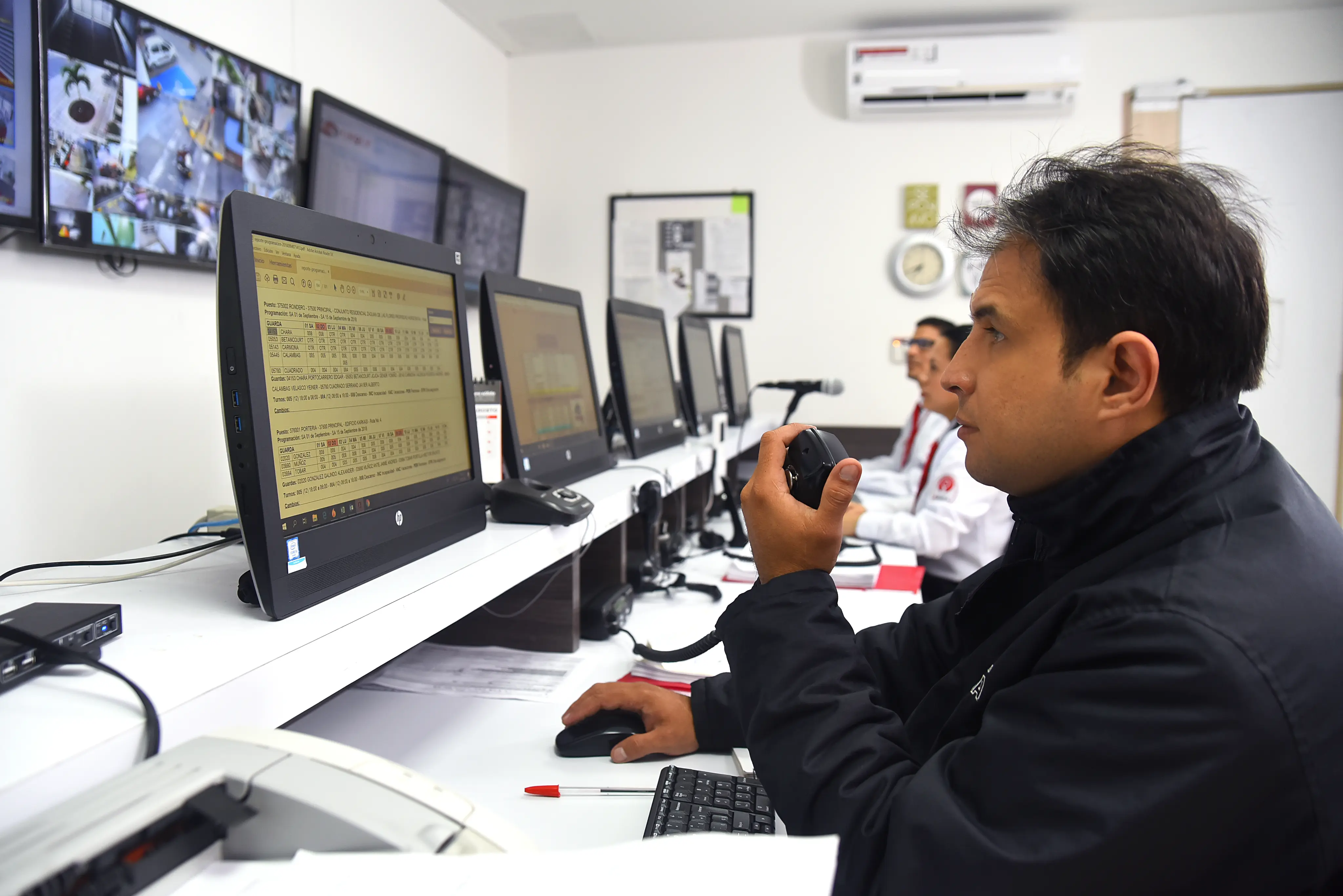 Man in a control room speaking into a handheld radio while monitoring multiple computer screens and security camera footage.