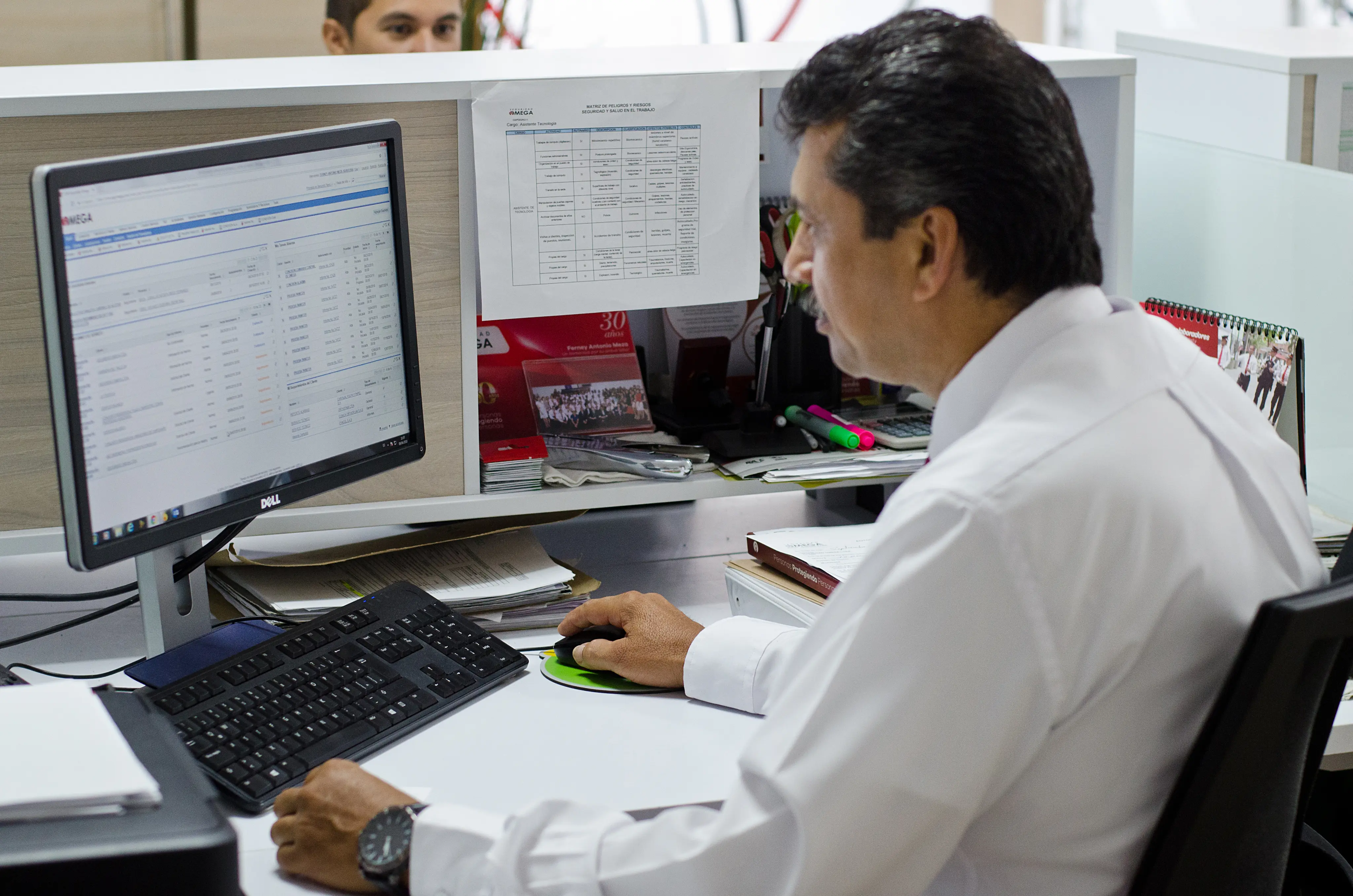 Man in a white shirt using a Dell computer at a cluttered office desk with documents, highlighters, and a calendar.