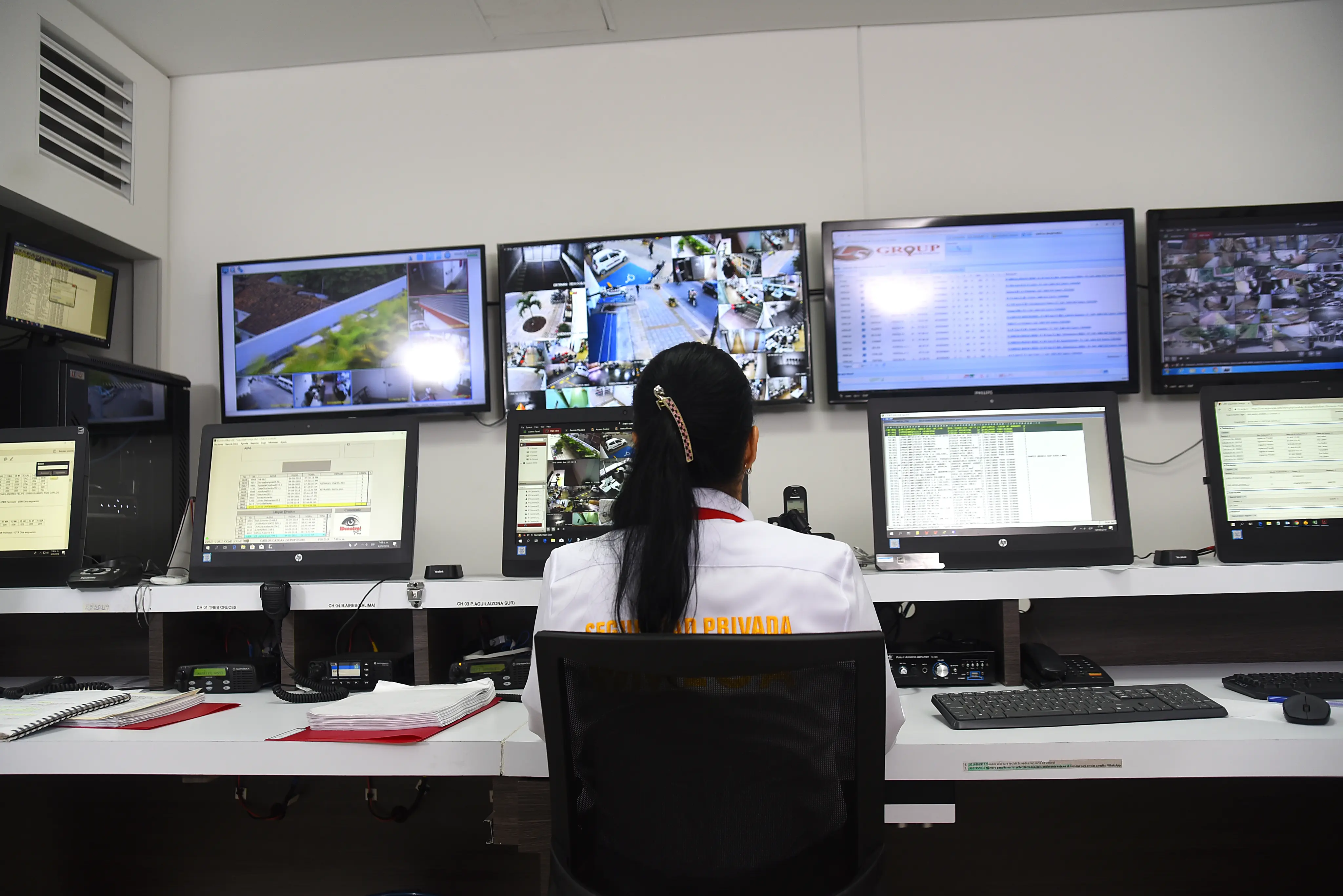 Security guard monitoring multiple surveillance camera feeds and data on several computer screens in a control room.
