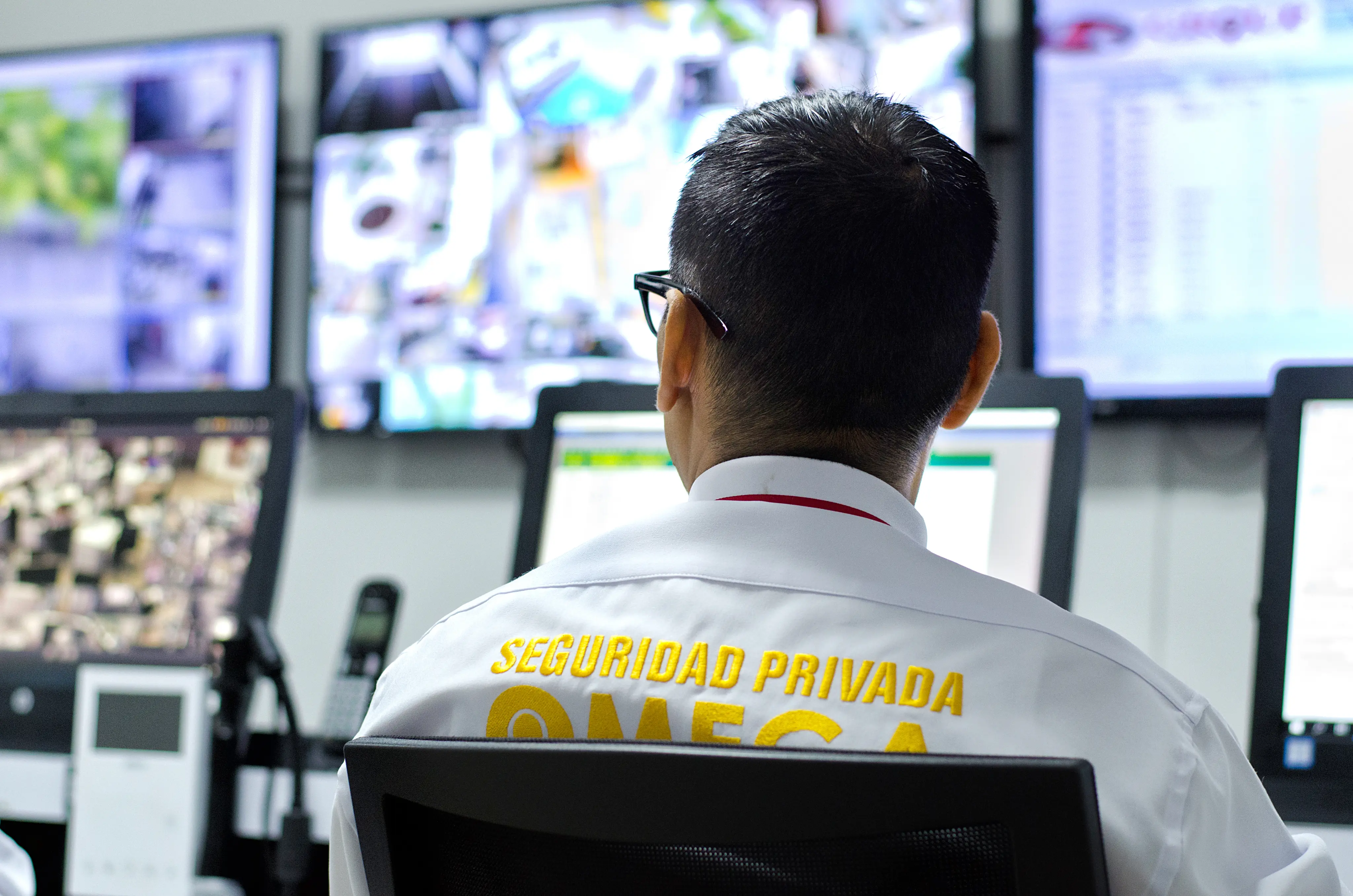 Man wearing a white shirt with 'Seguridad Privada OMEGA' watching multiple security monitors.
