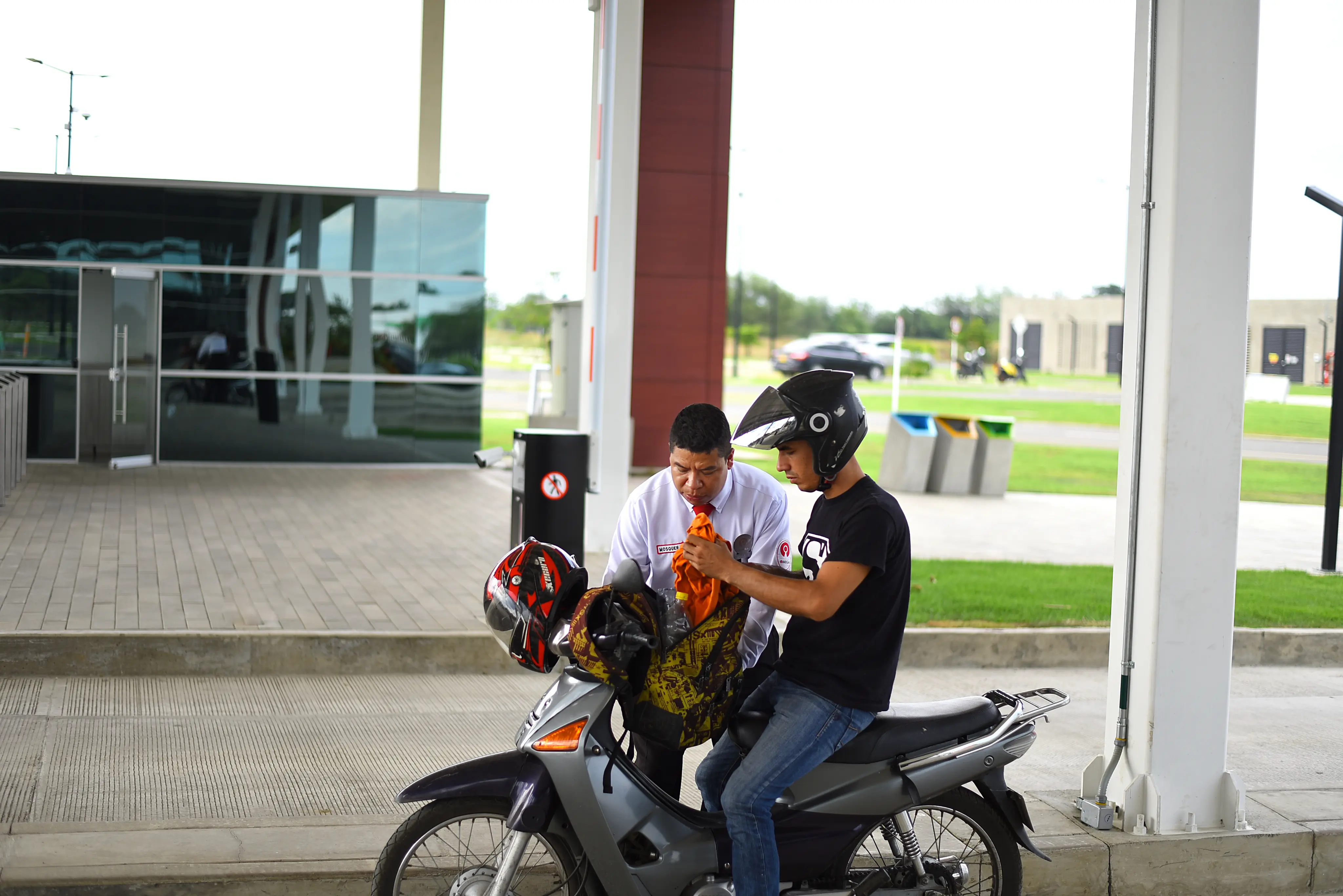 Man wearing a black helmet sits on a motorcycle while another man in a white shirt looks at an orange item they are holding together.
