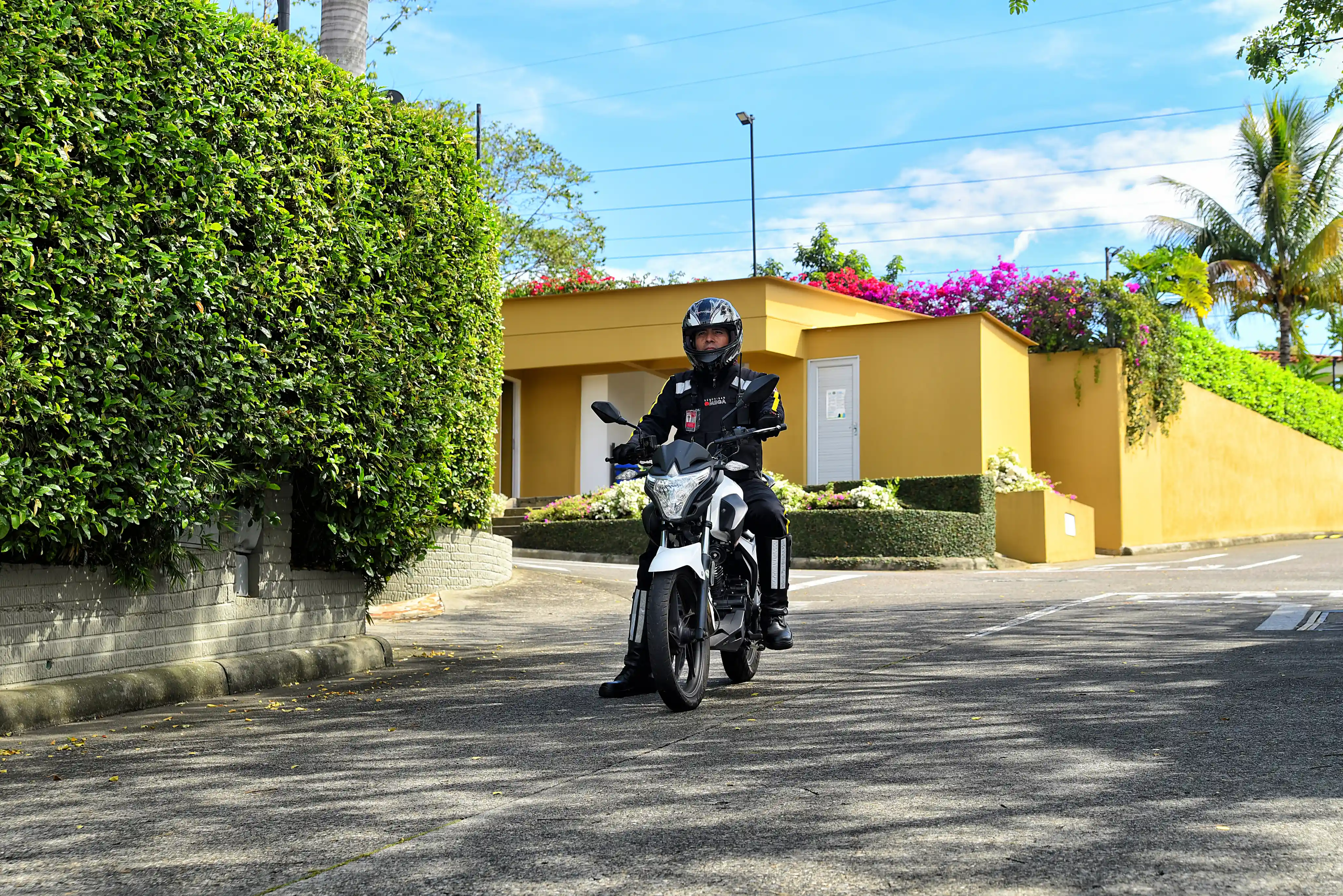 Motorcycle police officer in black uniform and helmet riding a white motorcycle on a sunlit street with green foliage and yellow buildings in the background.