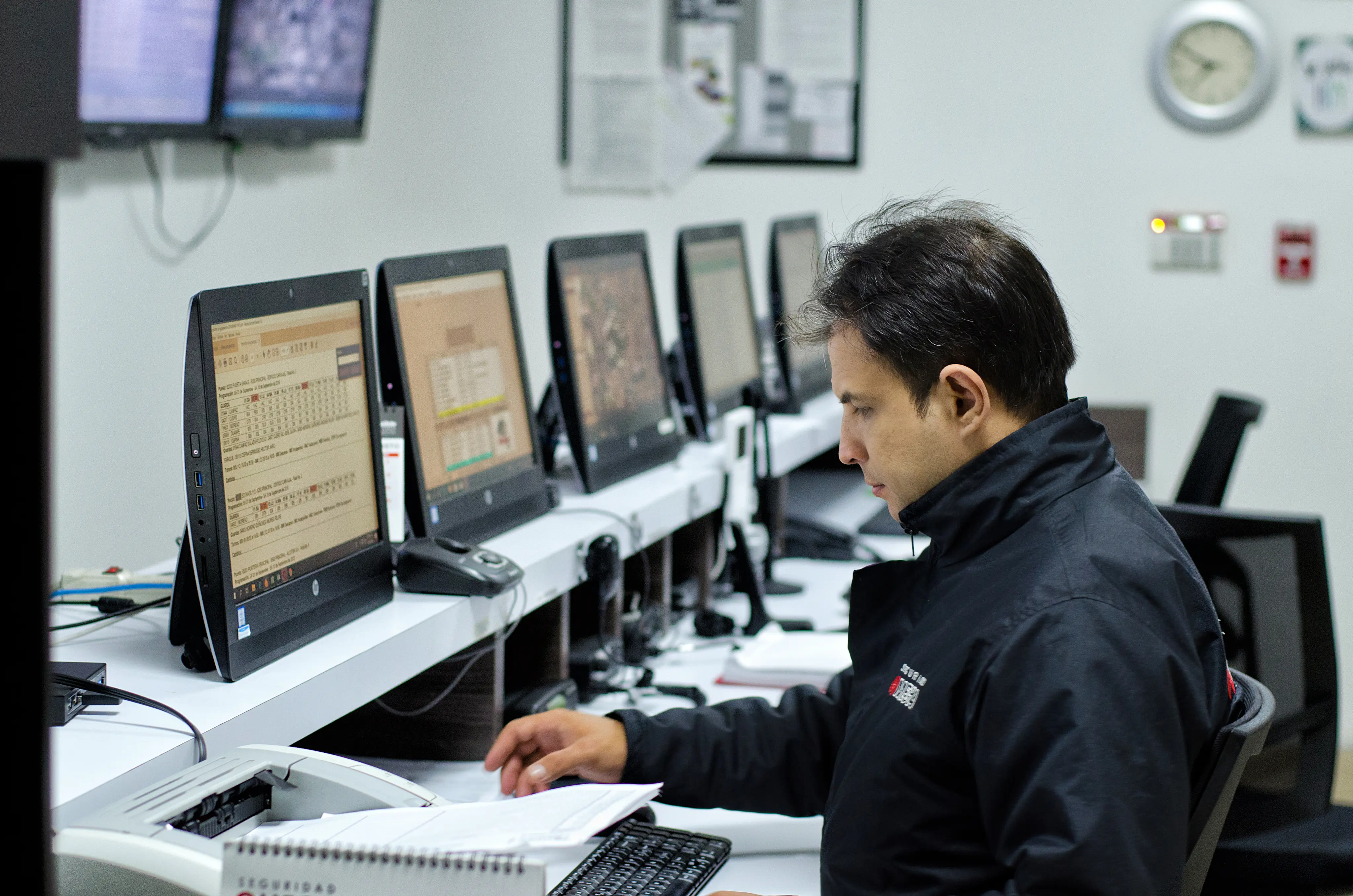 Man in a black jacket working at a row of computers in a control room.