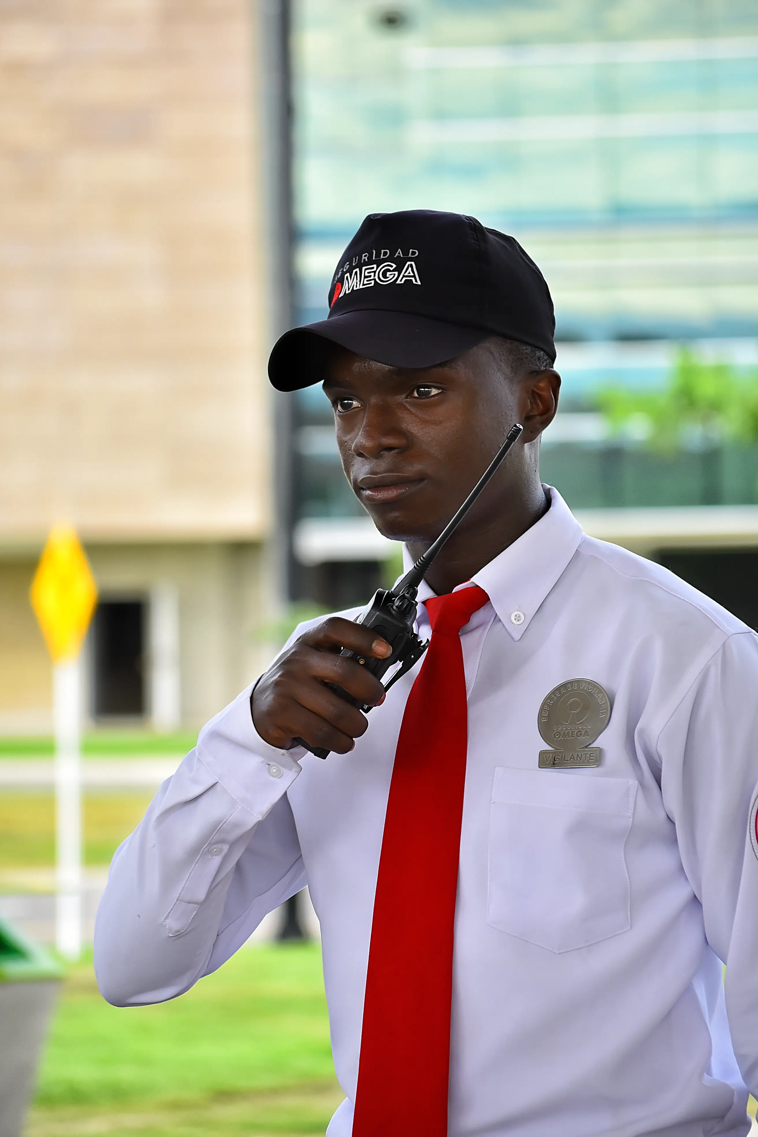 Security guard in white shirt, red tie, and black cap using a walkie-talkie outdoors.
