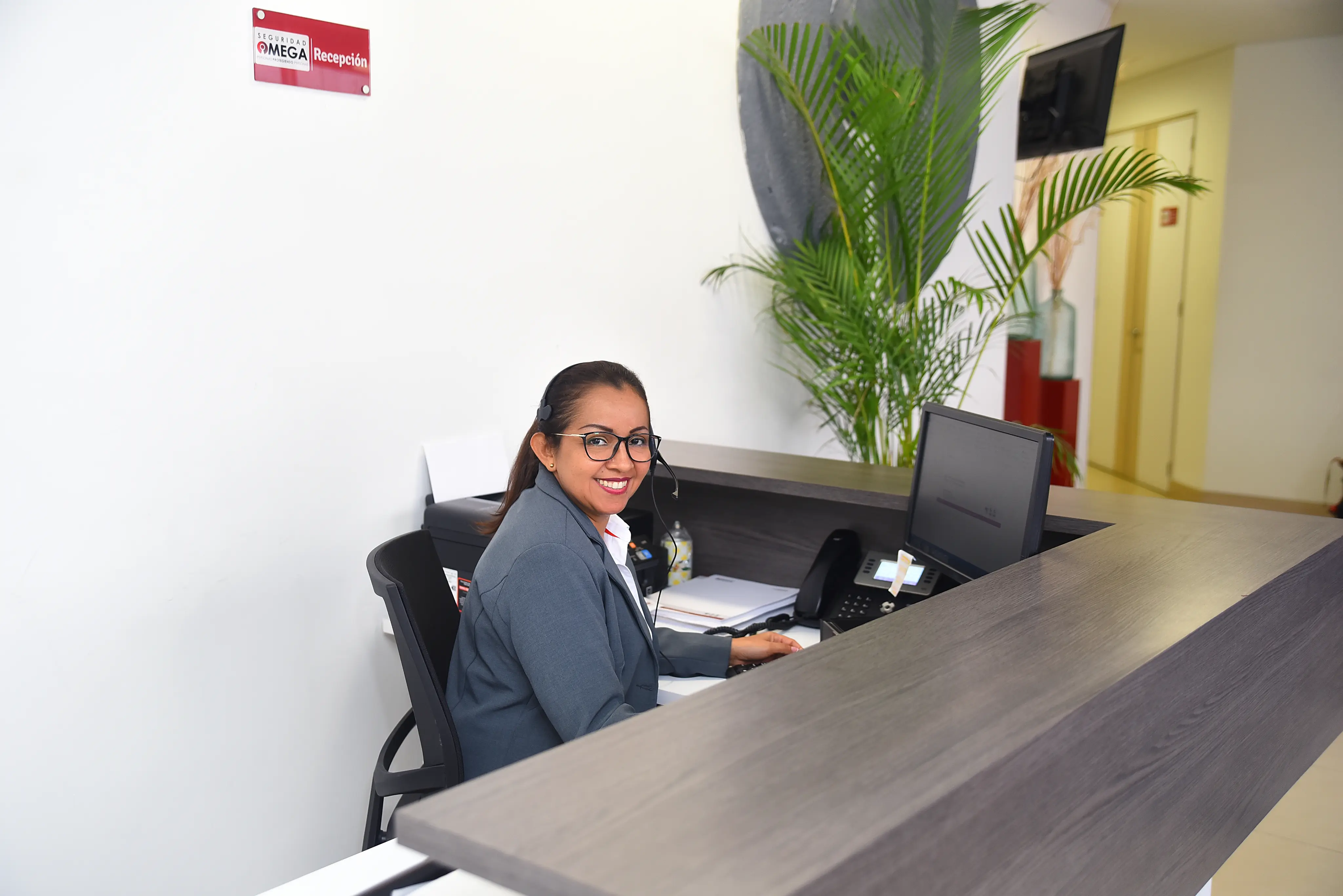 Smiling receptionist with glasses and headset sitting at a modern reception desk with computer and telephone, with a plant in the background.