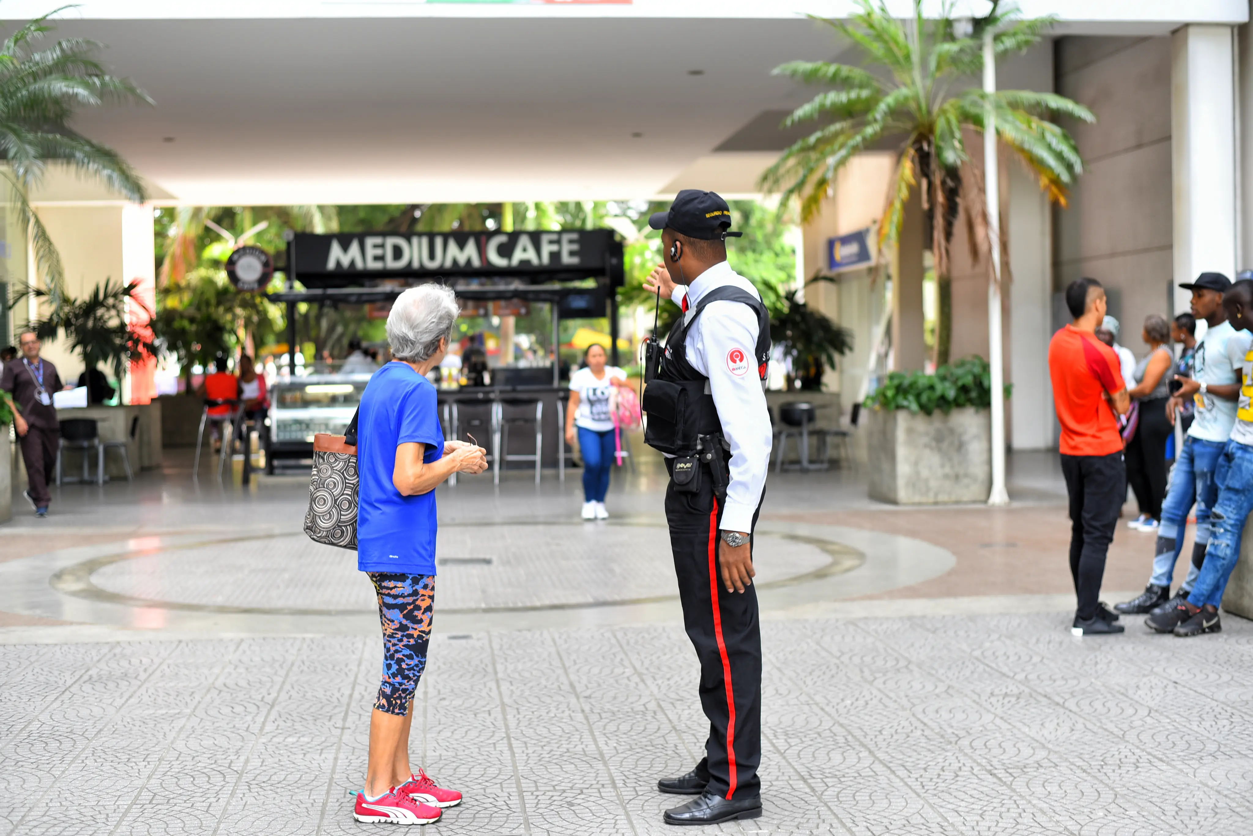 Security guard giving directions to a woman in athletic clothing outside a cafe in an outdoor public space.
