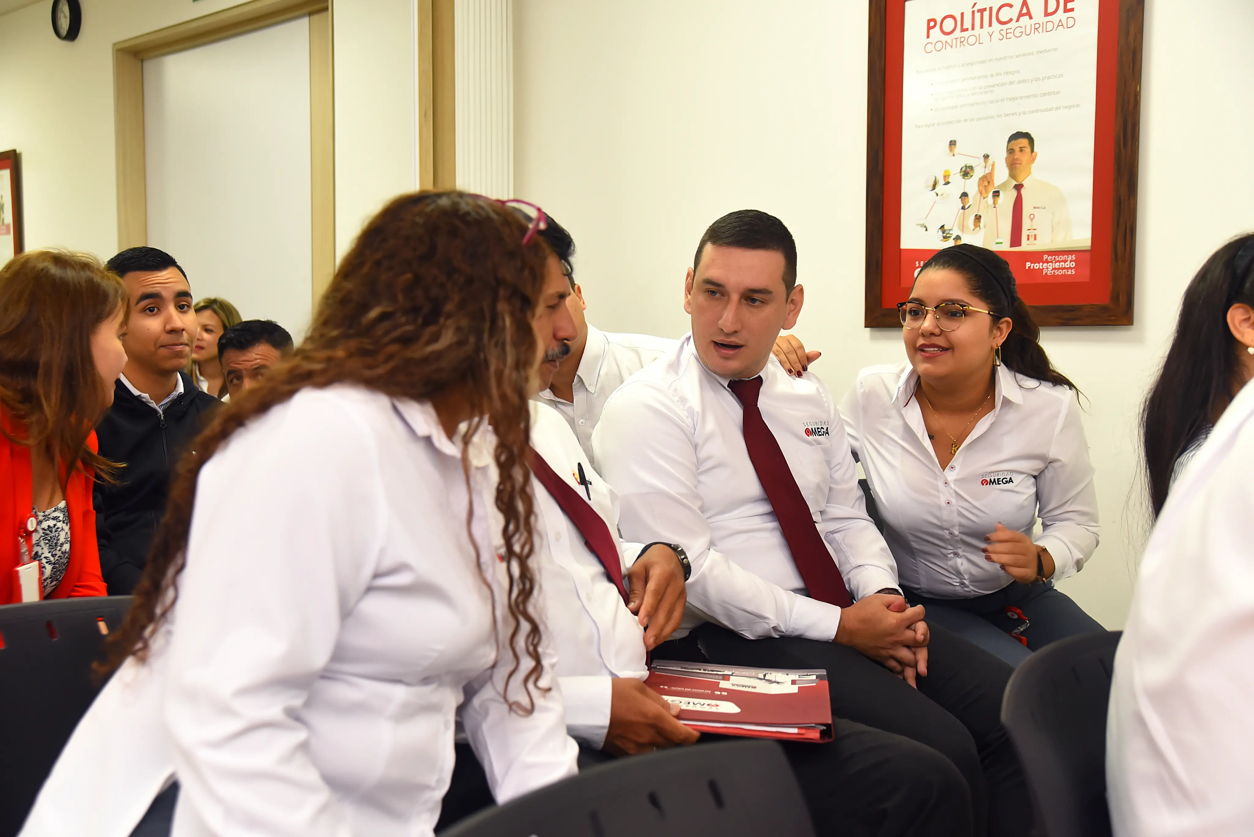 Group of employees in white shirts and red ties engaged in conversation in a meeting room with a policy poster on the wall.