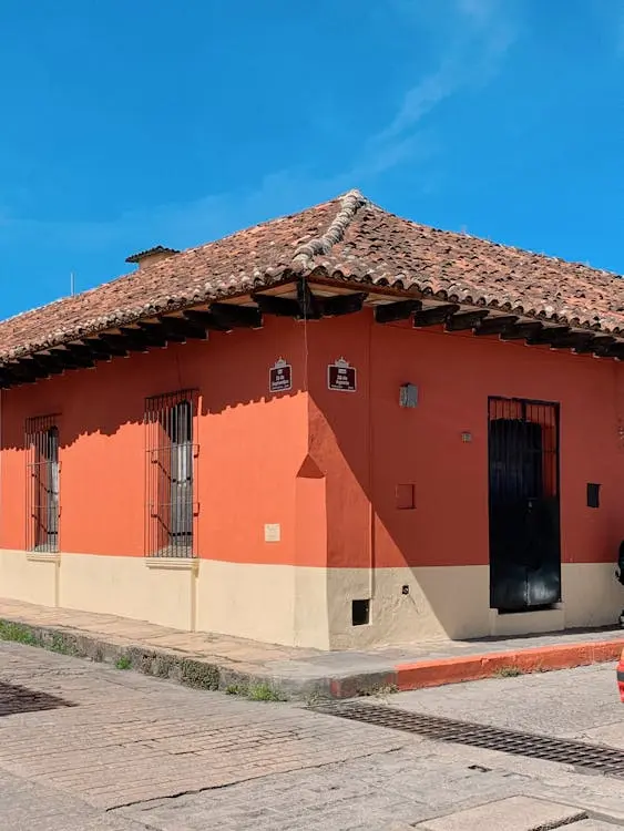 Corner view of a traditional single-story building with terracotta roof tiles, orange walls, barred windows, and a black door under a clear blue sky.