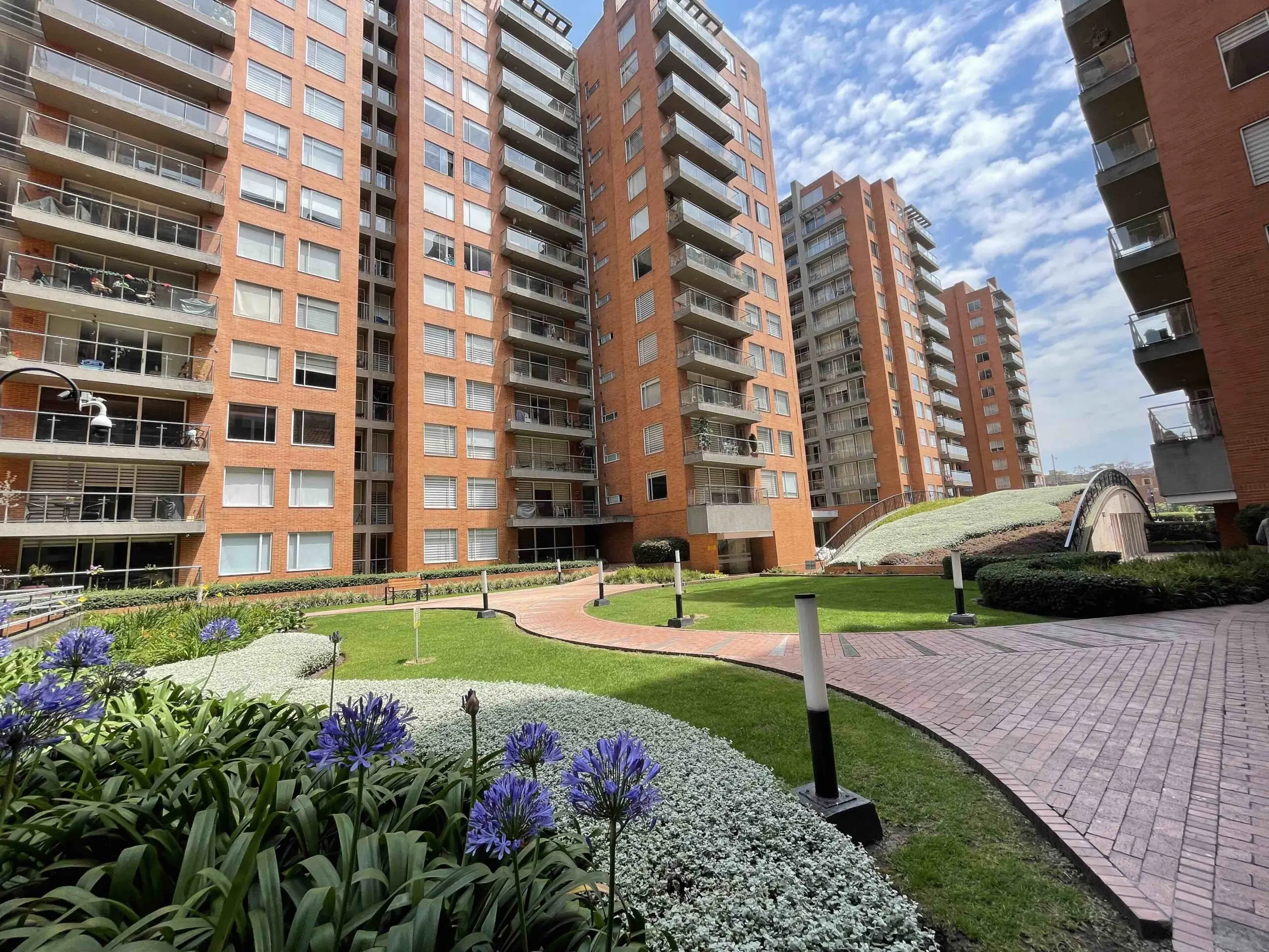 Brick apartment buildings surrounding a landscaped courtyard with green grass, flower beds, a curved brick pathway, and a small pedestrian bridge.