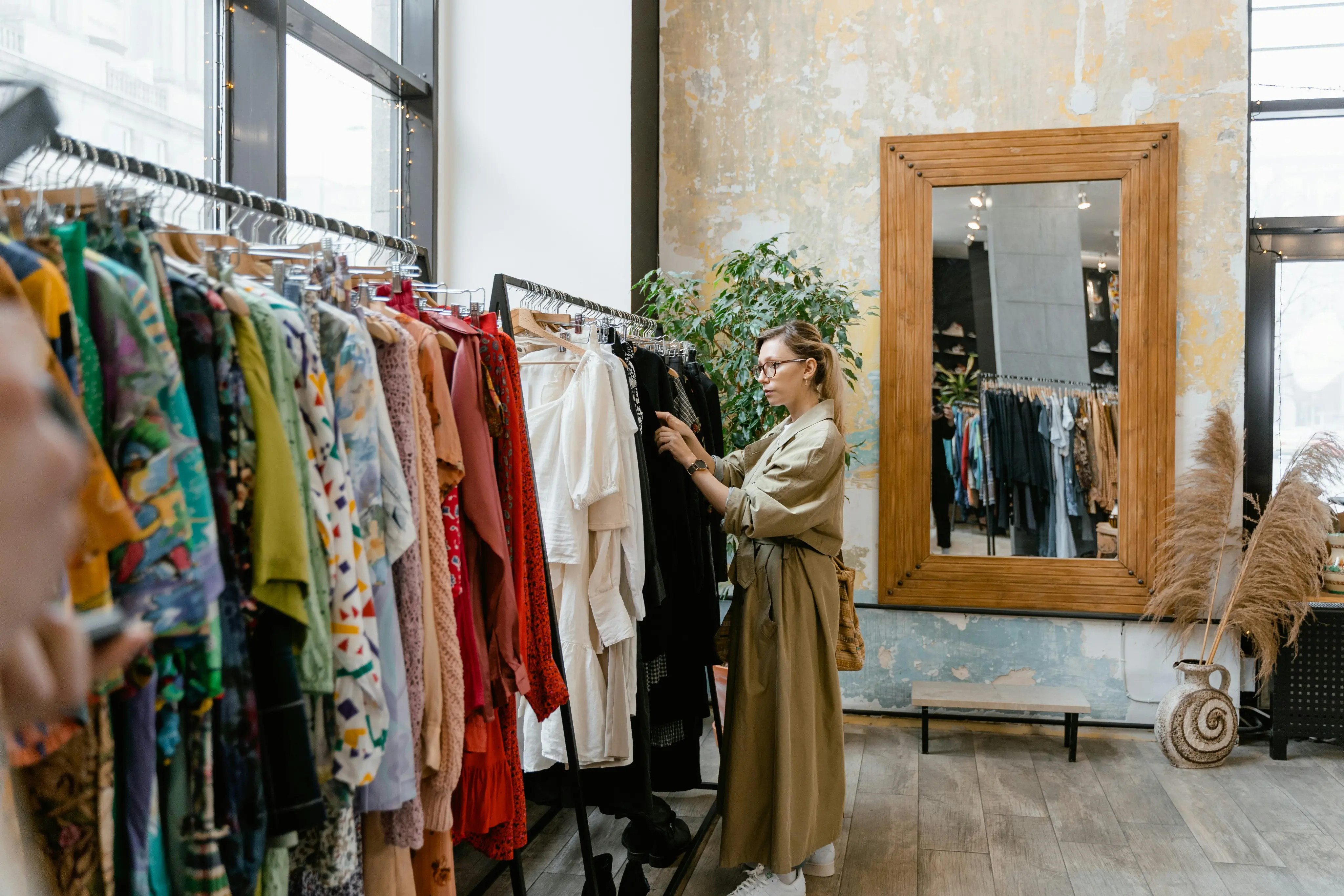 Woman wearing glasses and a long beige trench coat browsing clothes on a rack inside a boutique with a large wooden-framed mirror on a peeling plaster wall.