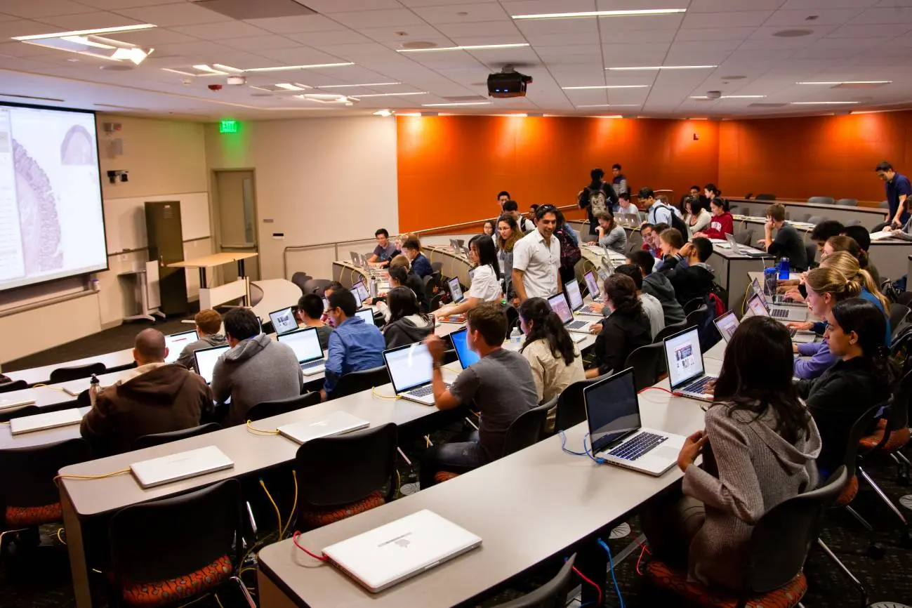 College classroom filled with students using laptops, with a professor engaging with them near the center.