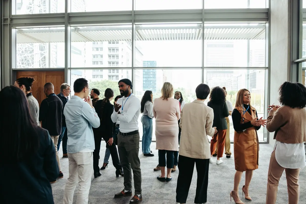 Group of diverse people engaged in conversations in a bright modern indoor space with large windows showing city buildings outside.