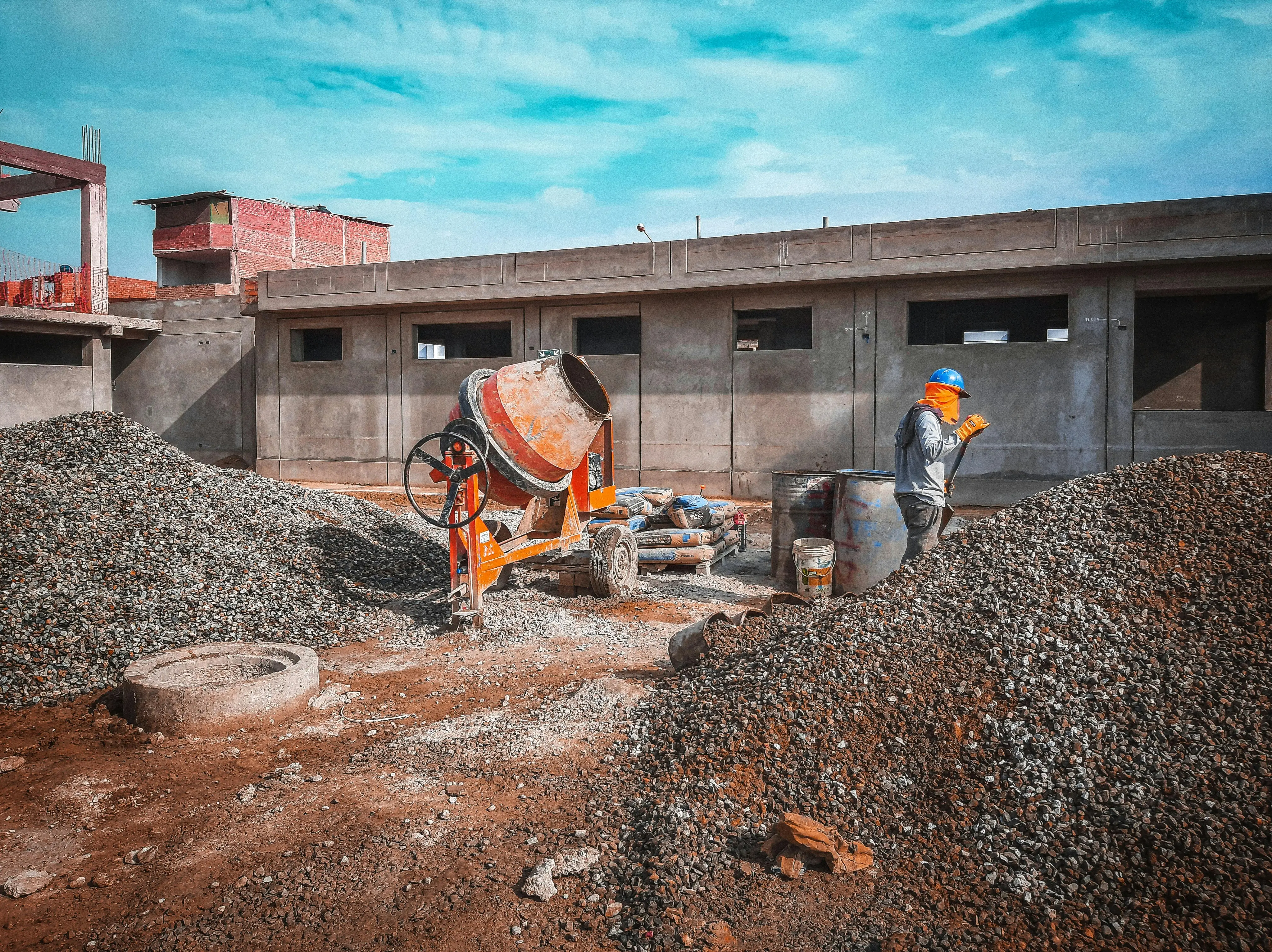 Construction site with a worker in a blue helmet and orange face cover standing near large piles of gravel and a concrete mixer.