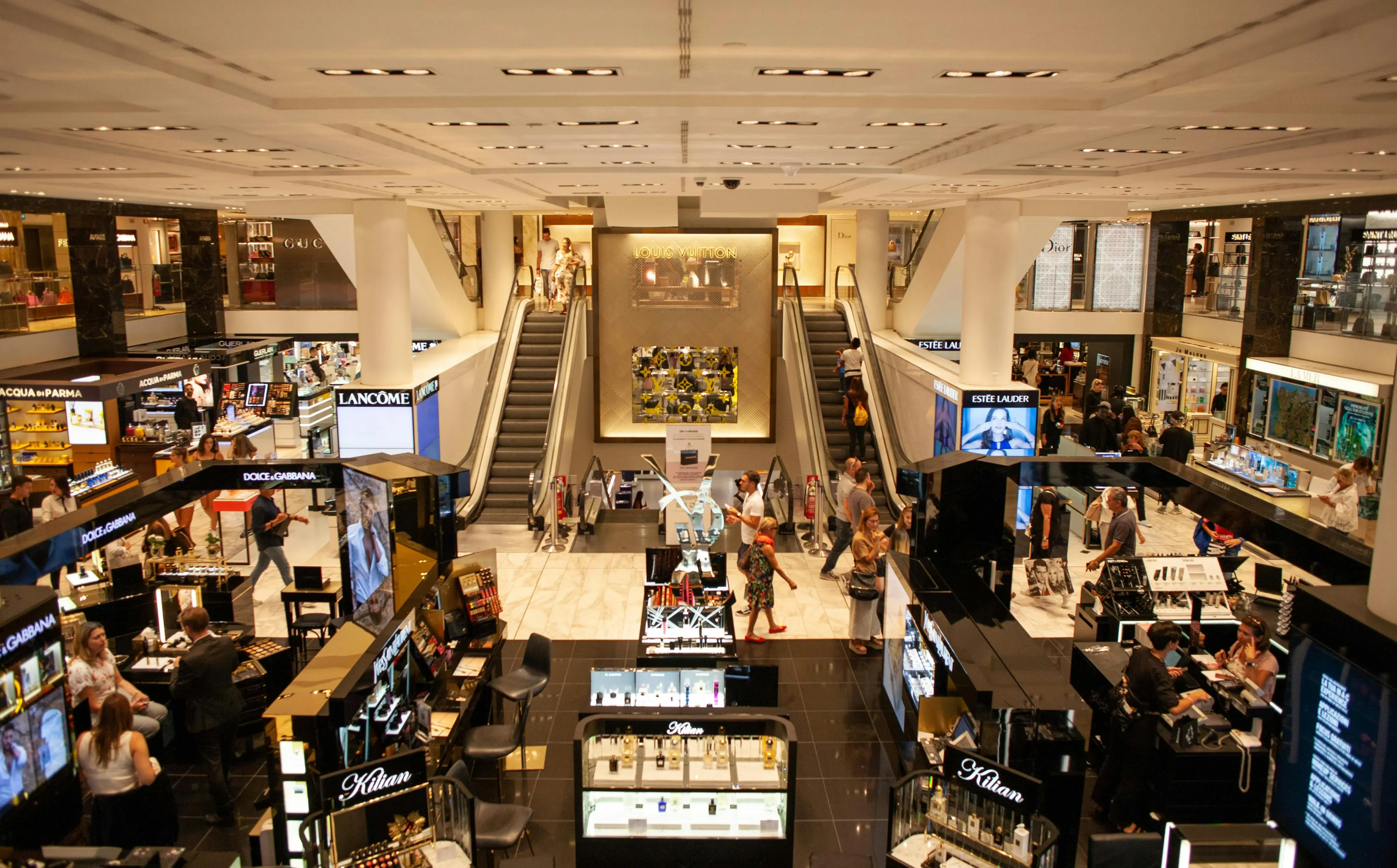 Interior view of a luxury department store with skincare and cosmetic counters, shoppers, and escalators leading to an upper level featuring a Louis Vuitton store.