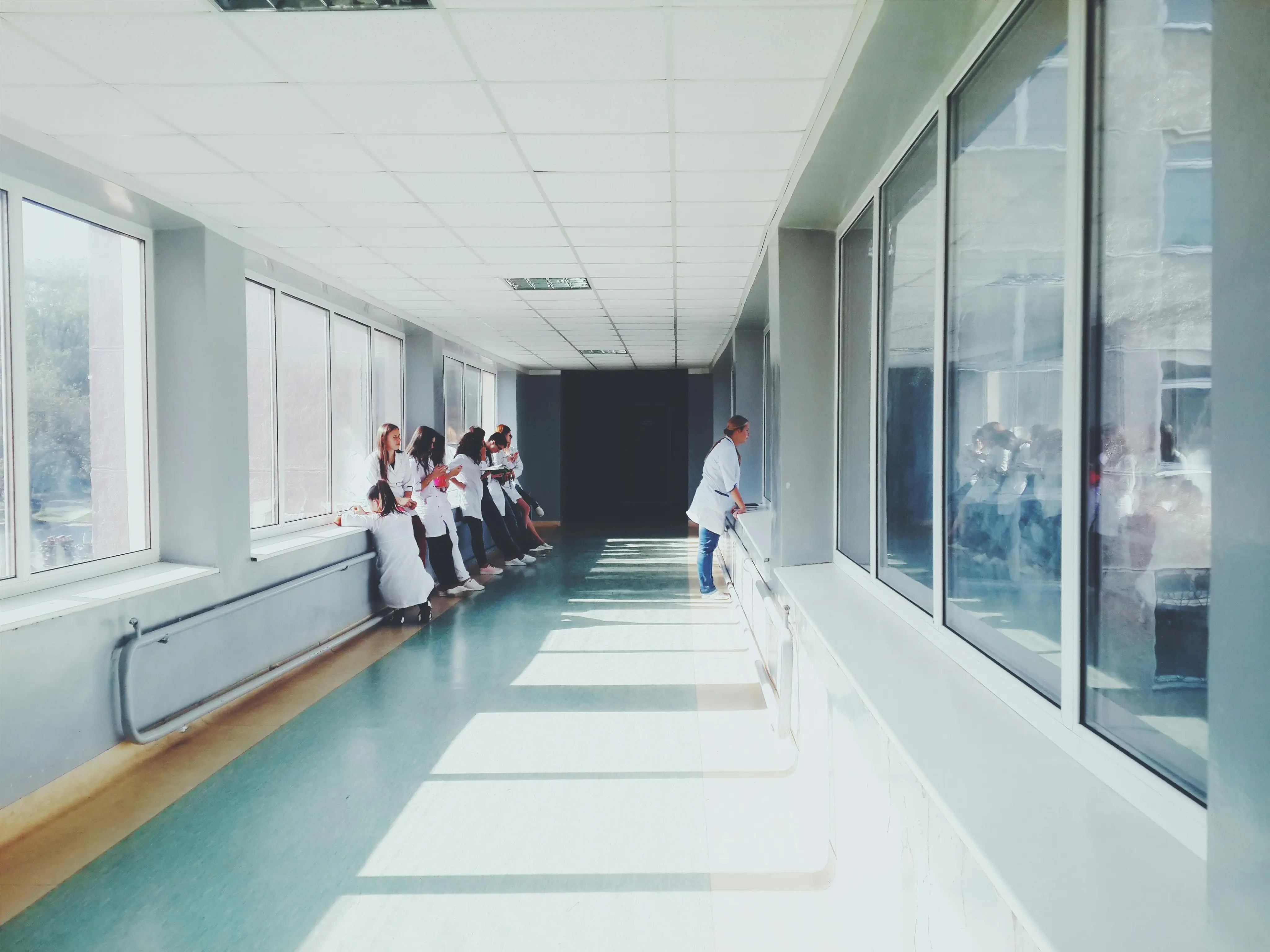Group of medical professionals in white coats standing and sitting along a sunlit hospital corridor with large windows.