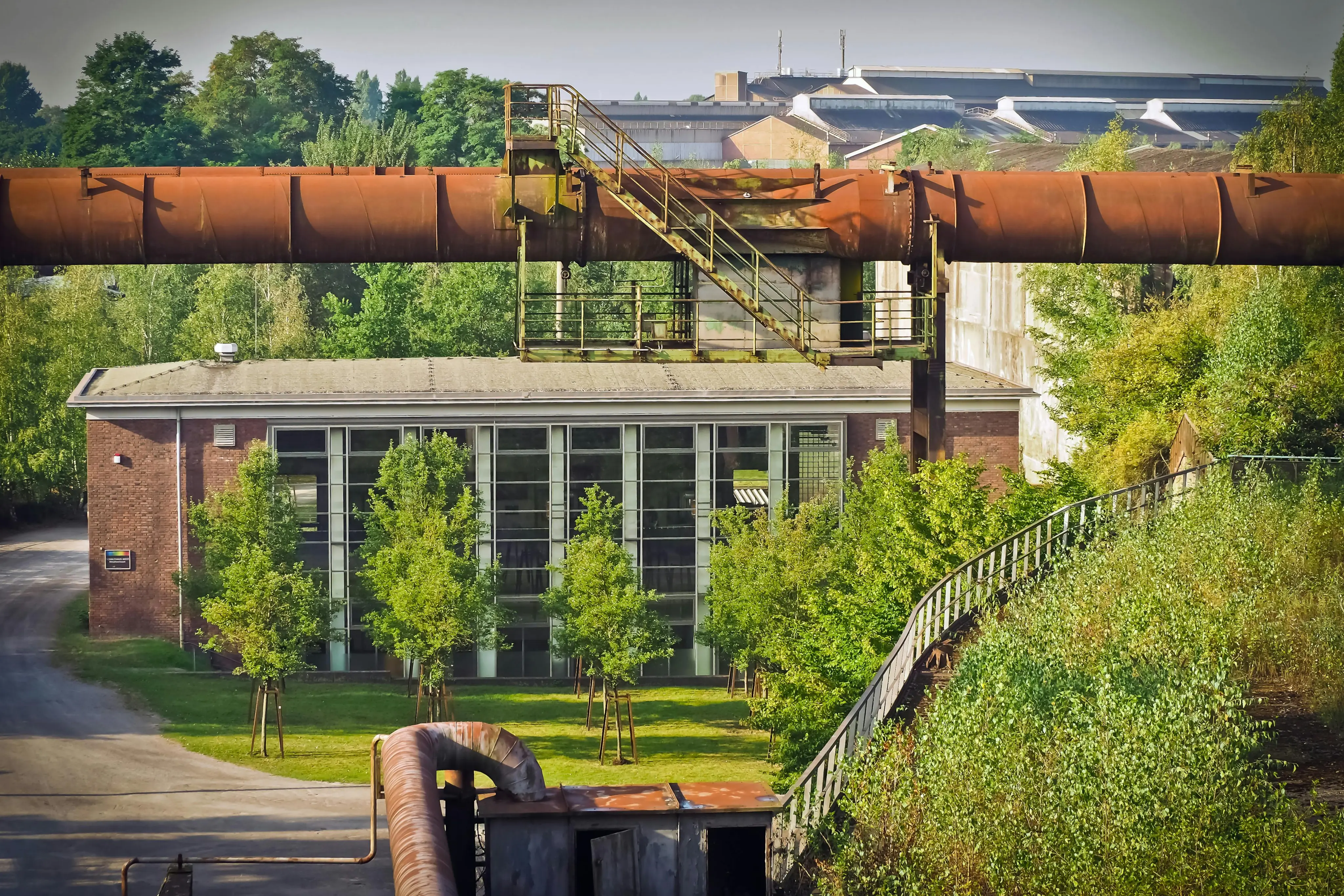Industrial site featuring large rusted ceiling pipe with staircase above a brick building surrounded by green trees and vegetation.