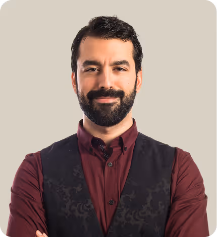 Portrait of a smiling man with dark hair and beard, wearing a maroon shirt and a black vest, standing with arms crossed against a plain background.