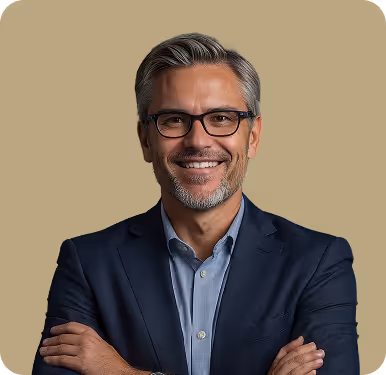 Smiling middle-aged man with gray hair and glasses wearing a navy blazer and blue shirt, standing with arms crossed against a beige background.