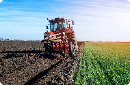 Red tractor plowing a field, turning dark soil next to a green crop under a blue sky.