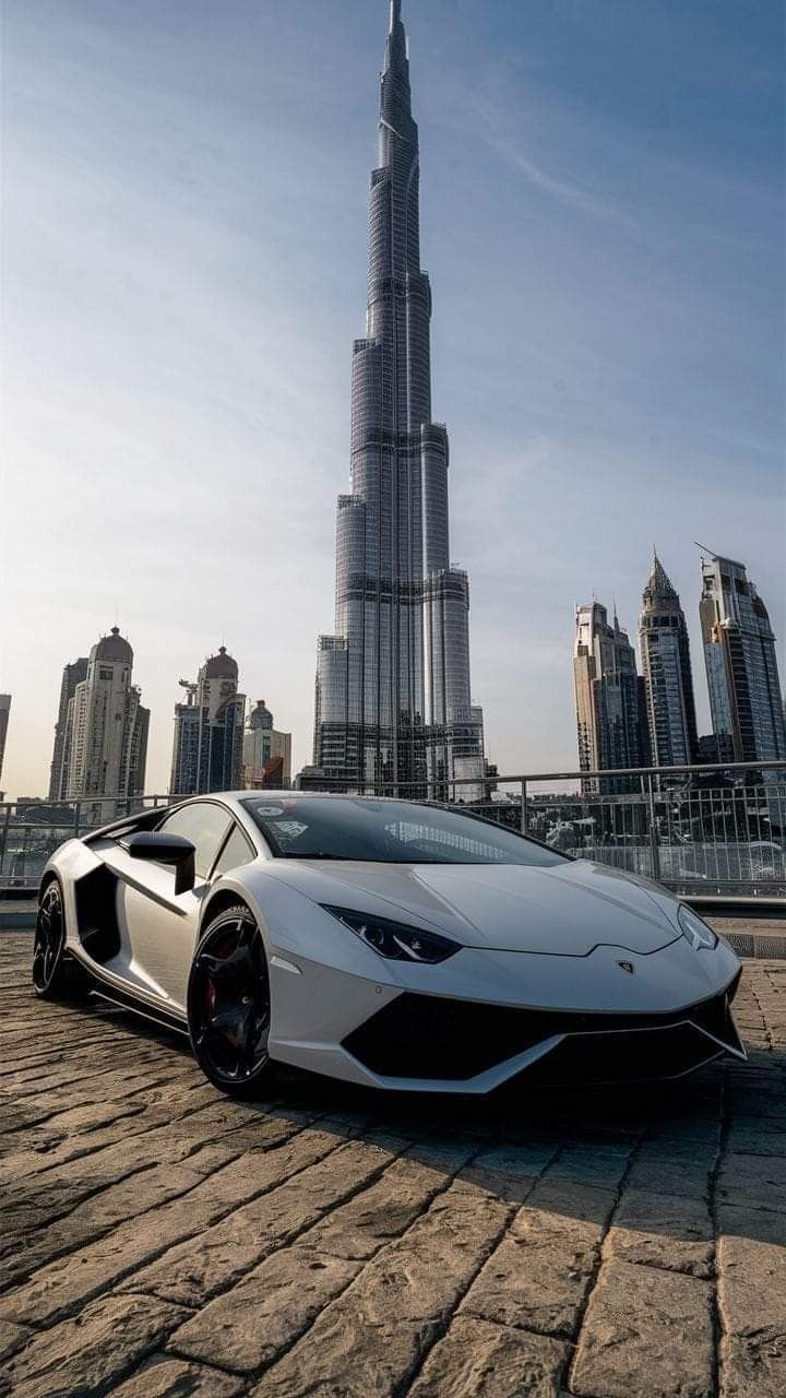 White Lamborghini parked on cobblestone pavement with the Burj Khalifa and Dubai skyline in the background under a clear sky.