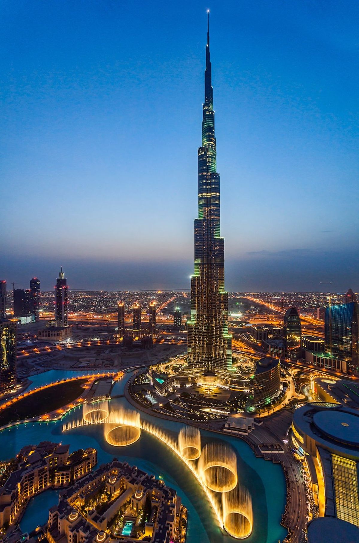 Illuminated Burj Khalifa skyscraper towering over Dubai Fountain and surrounding cityscape at dusk.