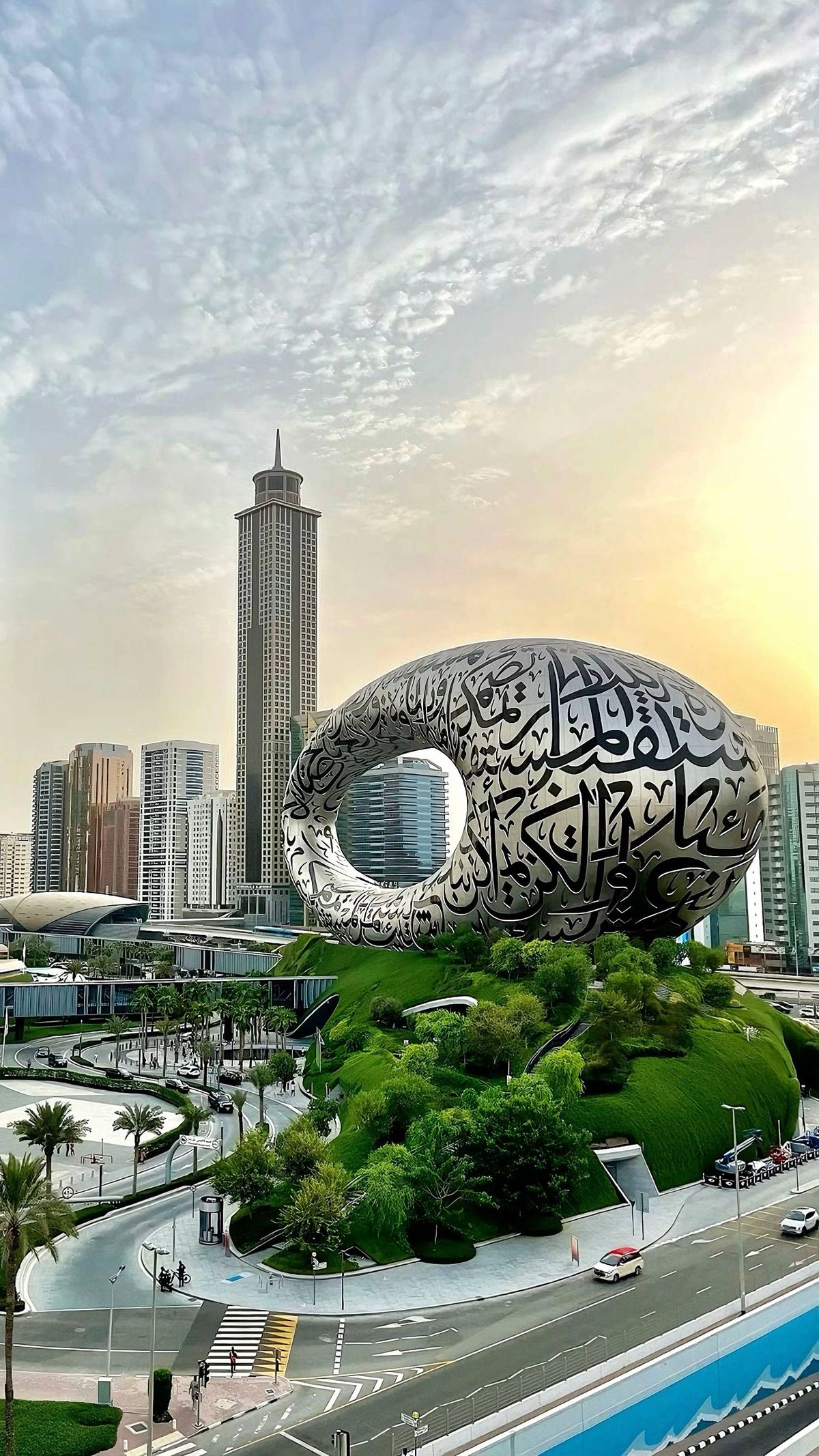 The Museum of the Future, a futuristic torus-shaped building with Arabic calligraphy, surrounded by greenery and city skyscrapers at sunset.