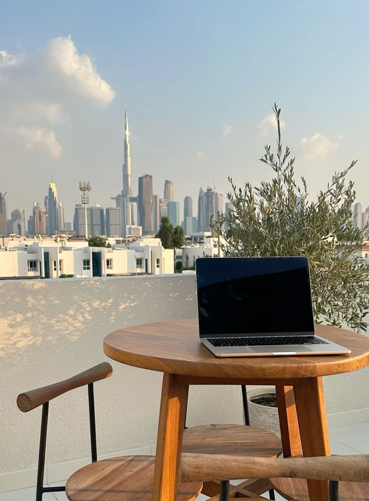 Laptop on a round wooden table with chairs on a balcony, overlooking a city skyline with skyscrapers including the Burj Khalifa under a clear sky.